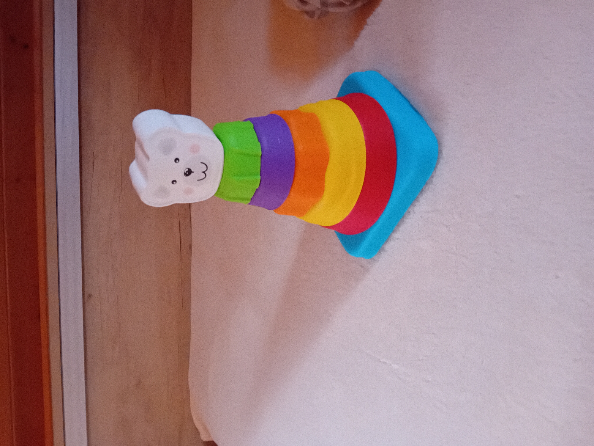 A brightly colored stacking toy is laid out on a textured, light-colored surface, likely a rug or carpet. The toy has a white, bear-shaped head with simple black eyes, a nose, and a pink blush on each cheek, along with a number "3" drawn on its face. Below the head, the toy features a series of stacked, colored rings in a rainbow pattern: green, purple, orange, yellow, and red. The base of the toy is a large, flat blue piece that flares outwards. The background is a mix of a wooden surface and the light-colored textured surface. The image is taken from a high angle, with the toy positioned diagonally across the frame. The scene appears to be indoors, with no visible signs of its exact location within Zuberec, Slovakia.