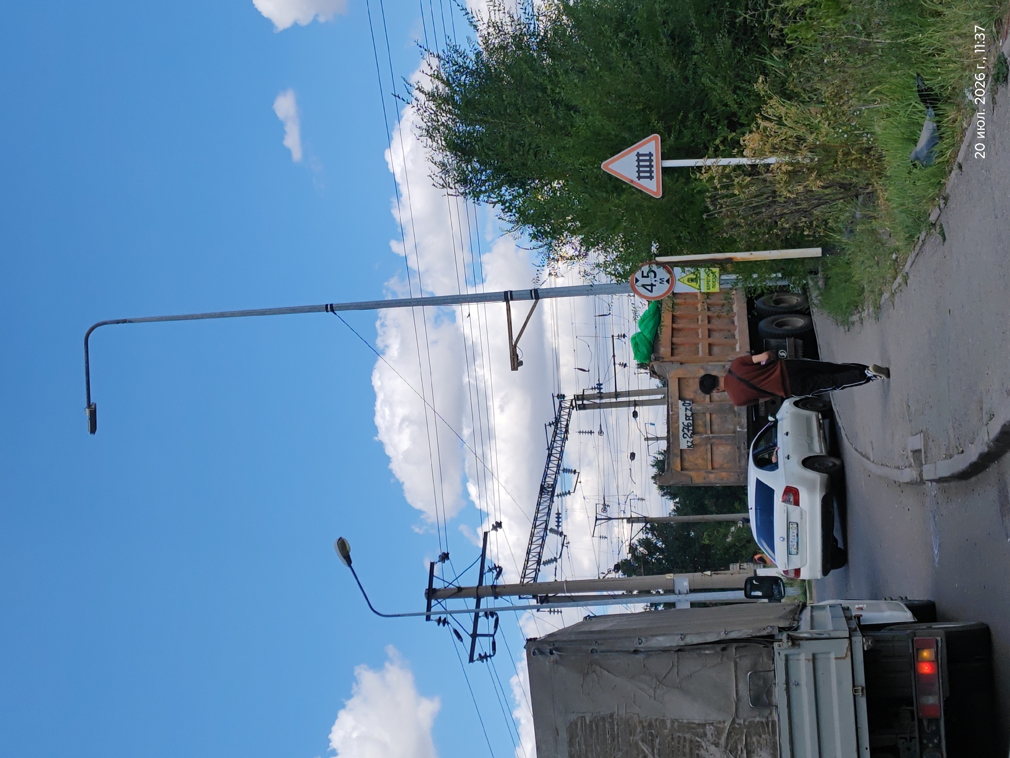 The image depicts a street scene in Astana, Kazakhstan, on July 20, 2026, at 11:37 AM. A young man in a maroon t-shirt and black pants is walking away from the camera on a paved sidewalk. To his left, a white sedan with license plate "C241AY 01" is driving away from the camera, and a large orange dump truck is parked on the right side of the road, facing away from the camera. A significant overhead electrical infrastructure system, likely for a railway or tram, is visible. In the background, trees and vegetation line the roadside. Two traffic signs are visible: one circular sign with "4.5 M" and a yellow warning sign below it, and a triangular sign with a railway crossing symbol. The sky is blue with scattered white clouds.