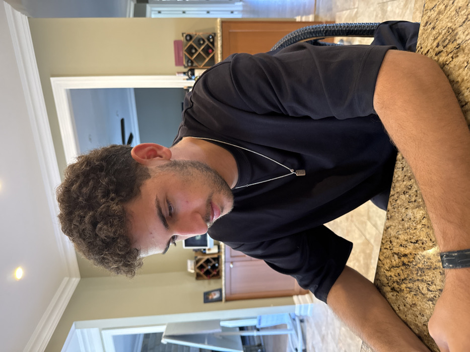 The image captures a young adult male seated at a speckled brown and gold kitchen island in a residential property located in Princeton, United States. The male, observed from the chest up, possesses dark, curly hair and light facial stubble. He wears a dark, short-sleeved t-shirt and a silver chain necklace with a small, rectangular pendant. His attention is directed downwards, suggesting engagement with an unseen object or task on the countertop. A dark wristband is visible on his left wrist. In the background, a domestic interior features light-toned walls, white crown molding, and recessed ceiling lights. A white, mobile easel is partially visible on the left. To the right, a wooden wine rack containing multiple bottles of red wine rests on wooden cabinetry. A doorway in the distance reveals an additional interior space equipped with a ceiling fan. The subject is seated in a chair with a dark, woven back, partially visible on the right.