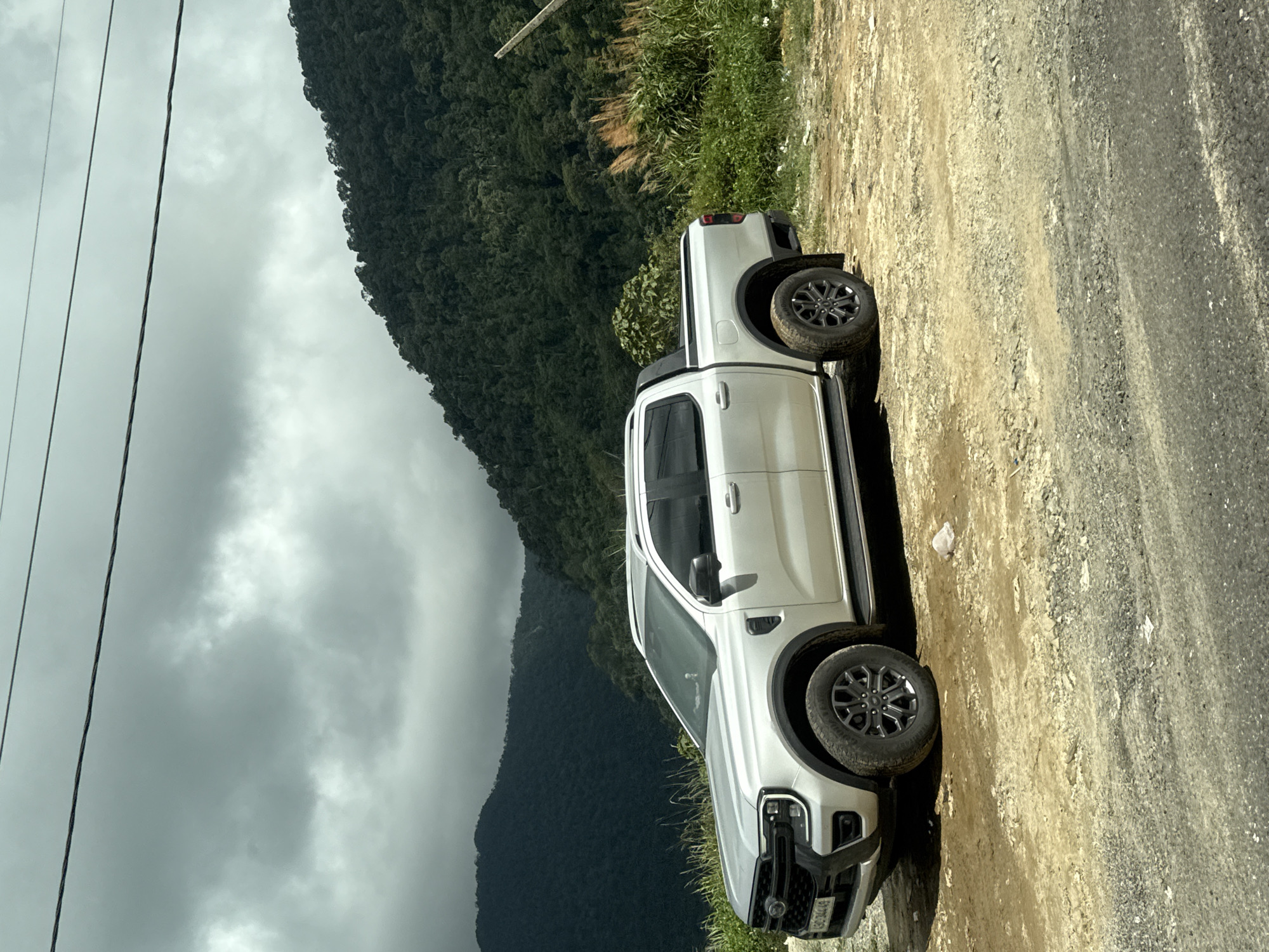 An overcast, cloudy sky looms over a mountainous, forested landscape. A silver pickup truck is parked on a dirt and gravel road. The truck is angled towards the left of the frame, with its front quarter visible. The road is uneven and appears to be in a rural or undeveloped area. Tall grasses and some sparse trees line the edge of the road on the right. The mountains in the background are covered in dense, dark green vegetation. The overall atmosphere is somber due to the cloudy weather.
