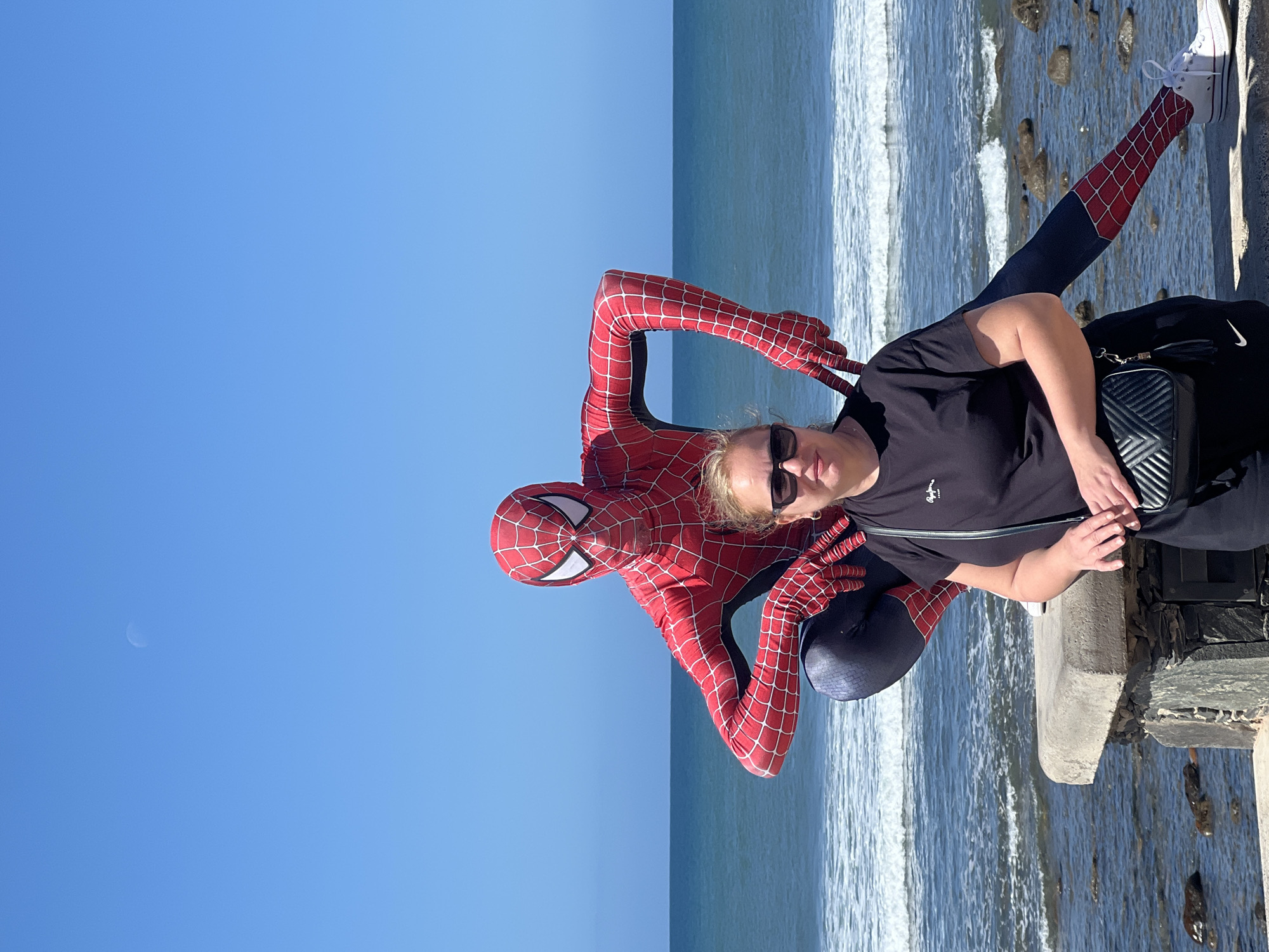 A woman and Spider-Man are posing for a photograph. The woman is wearing sunglasses, a black shirt, and black pants, and she is holding a black purse. Spider-Man is wearing his red and blue costume. They are standing on a concrete structure next to the ocean, with rocks visible in the water. The background features a clear blue sky and the ocean. The image appears to have been taken in San Bartolomé de Tirajana, Spain, given the coastal environment and the specific architectural features along the coastline, which aligns with typical structures found in the area.