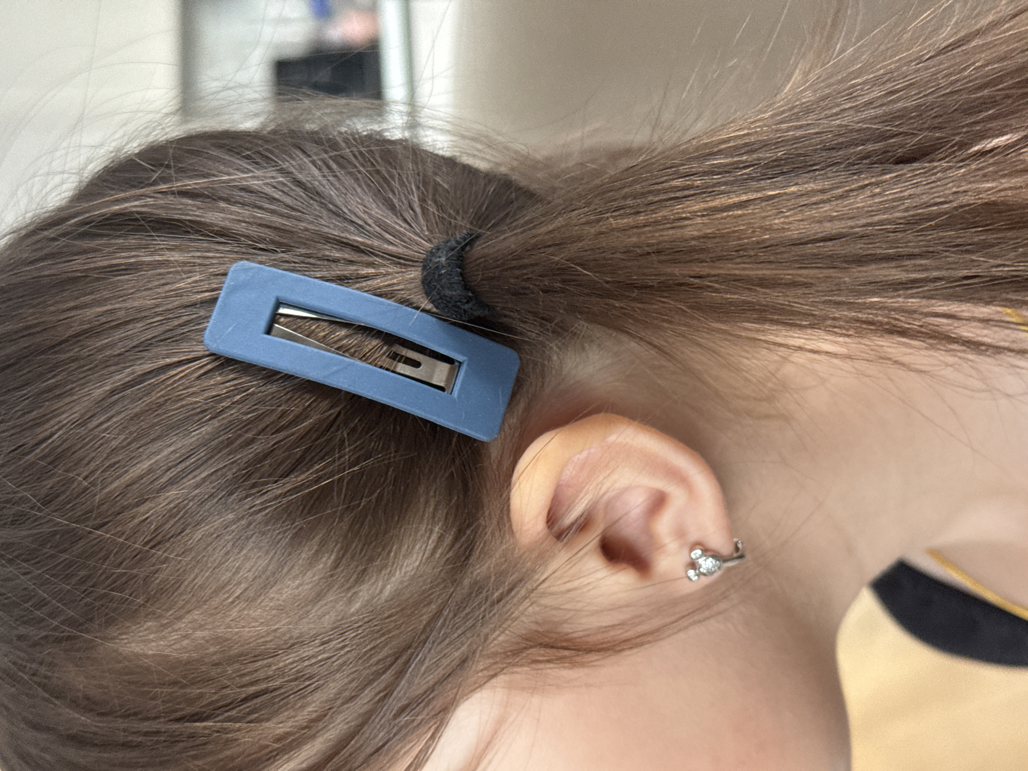The image provides a close-up, side-profile view of a person's head, focusing on their hair and ear. The person has light brown hair, which is gathered into a ponytail secured by a black, textured elastic hair tie. A matte blue, rectangular snap hair clip (barrette) is positioned on the side of the head, holding some hair.
The person's right ear is visible, featuring a small, silver-toned earring in the lobe. The earring appears to be shaped like a stylized animal, possibly a mouse, with small clear stones. A sliver of yellow-gold fabric, likely part of a garment, is visible near the neck, adjacent to a darker, possibly black, fabric.
The background is softly blurred, suggesting an indoor setting with indistinct light-colored features. The activity depicted appears to be personal grooming or a casual moment. There is no visible text within the image. The time of day is not explicitly indicated, and no specific location cues for Murom, Russia, are present beyond the indoor setting. Emotions cannot be determined as no facial features are visible.