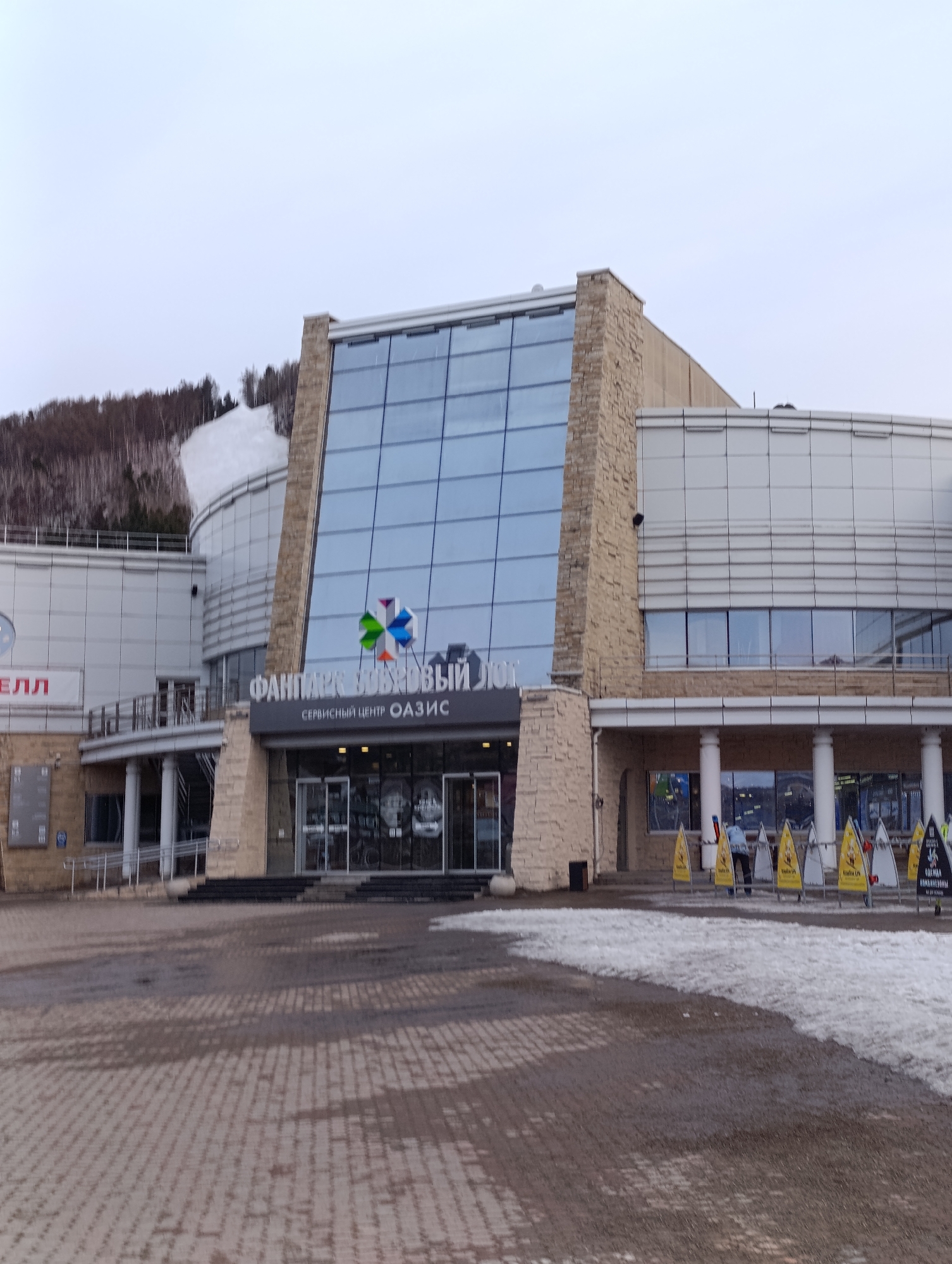 The image shows the facade of the Funpark Bobrovy Log service center in Krasnoyarsk, Russia, on an overcast day. The building has a central glass panel section flanked by light-colored stone structures. The entrance is a double set of glass doors with signage above it in Russian. A paved brick plaza leads to the building entrance. Patches of melting snow are visible on the plaza. Yellow A-frame signs stand in front of the building, likely advertising the park. There are people visible on the plaza in front of the building. In the background, a snow-covered slope and forested area are visible behind the building.