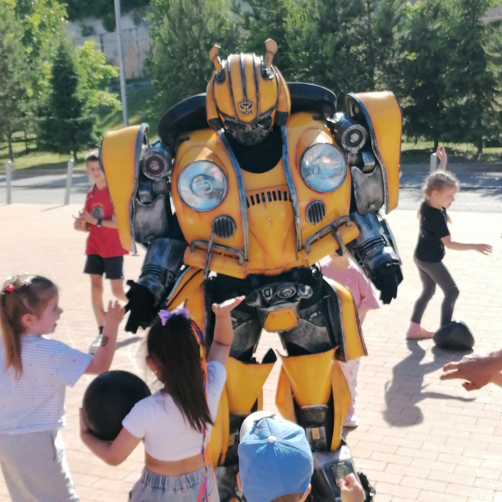 A large, yellow and grey costume resembling the Transformer character Bumblebee stands centrally on a paved outdoor surface, facing slightly left. The costume features circular headlights on its chest, a prominent Autobot symbol on its helmet, and antenna-like structures on top. Several children are gathered around it.
In the left foreground, a young girl with light hair, wearing a striped white top and grey pants, looks up at the costume with her left hand raised. She holds a black ball in her right hand. To her right, another young girl with dark hair, wearing a white crop top and denim shorts, also raises her left hand towards the costume while holding a black ball in her right. Partially visible at the bottom, a young boy in a blue baseball cap looks upwards.
To the left of the costume, a boy in a red t-shirt and black shorts stands, observing the scene. To the right of the costume, a girl in a black t-shirt and dark leggings stands with her arms slightly raised, with a black ball near her feet. A partial adult hand is visible in the lower right, pointing towards the girl and the ball.
The ground is paved with light-colored tiles, exhibiting distinct shadows from the figures, indicating bright daylight. The background features lush green trees and foliage, characteristic of an urban park or public garden in the city of Трубачеевка, Russia.