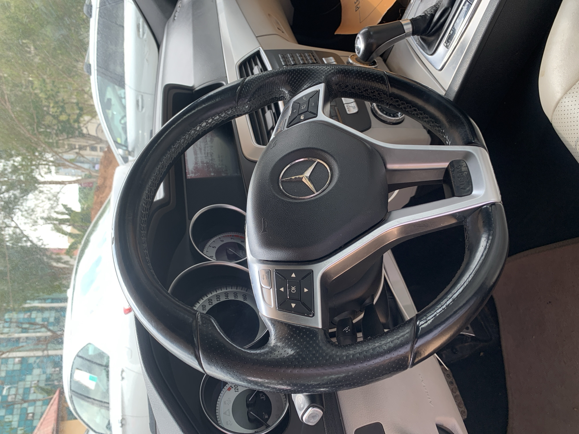 The media file displays the interior of a Mercedes-Benz car, rotated 90 degrees clockwise from its typical upright orientation. No people are visible.
The central focus is a black leather steering wheel with perforated grips on the sides and silver-colored spokes. A prominent Mercedes-Benz three-pointed star logo is centered on the airbag cover. The left spoke of the steering wheel features a cluster of multifunction buttons, including volume controls, menu navigation arrows, and an "OK" button. The text "AIRBAG" is visible at the bottom of the steering wheel's center hub.
Behind the steering wheel, portions of the instrument cluster are visible, displaying analog gauges with white markings on a black background, including a speedometer marked up to at least 160, a tachometer, and smaller fuel and temperature gauges. The dashboard incorporates silver trim elements and air vents. To the right, an automatic gear shifter is seen on the center console. A light-colored (possibly off-white or beige) perforated leather car seat is partially visible on the far right.
Through the windshield, a daytime scene is present, characterized by diffused lighting, suggesting overcast or cloudy weather. The exterior view includes part of a white vehicle, green trees, and architectural details of buildings in the background, indicating an urban or suburban setting. The car's interior appears to be clean and well-maintained, suggesting it is either parked or stationary.