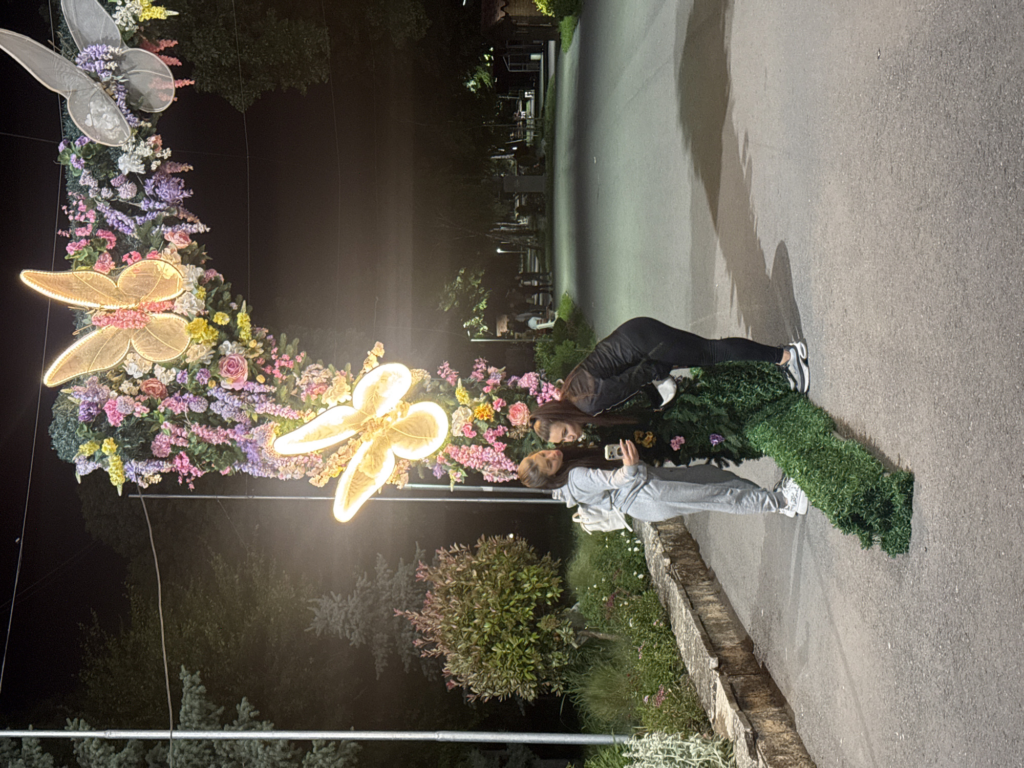 Two young women are positioned in front of an illuminated floral display adorned with decorative butterflies in Shumen, Bulgaria. The woman closer to the foreground is wearing a light gray sweatsuit and a white backpack; she is holding a phone and appears to be taking a photo. The other woman is wearing a dark tracksuit. The floral display is arranged in a semi-circular shape, with a mix of flowers in various colors. Behind the women and the display, there are bushes and trees. In the background, there is a paved path or road, along with streetlights and buildings. The image seems to be taken at night.