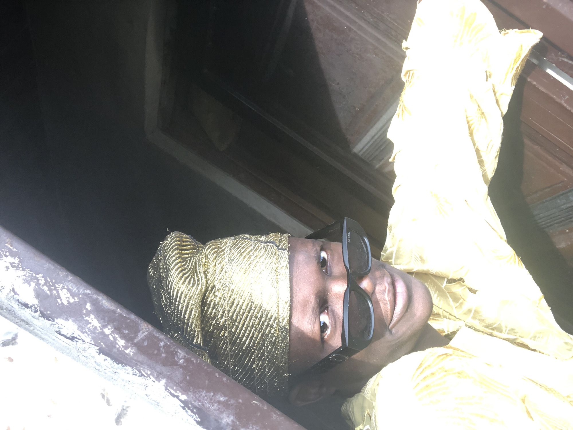 The image shows a close-up, selfie-style shot of a dark-skinned person wearing a gold, textured cap and black sunglasses. They are also wearing a yellow, patterned garment. The person is looking directly at the camera with a slight smile. The background is dark and appears to be an interior space with some wooden elements and overhead structures, possibly a doorway or ceiling. The lighting is strong, casting shadows and highlighting the textures of the cap and garment. The context of Ado, Nigeria, is not visually evident in the image itself.