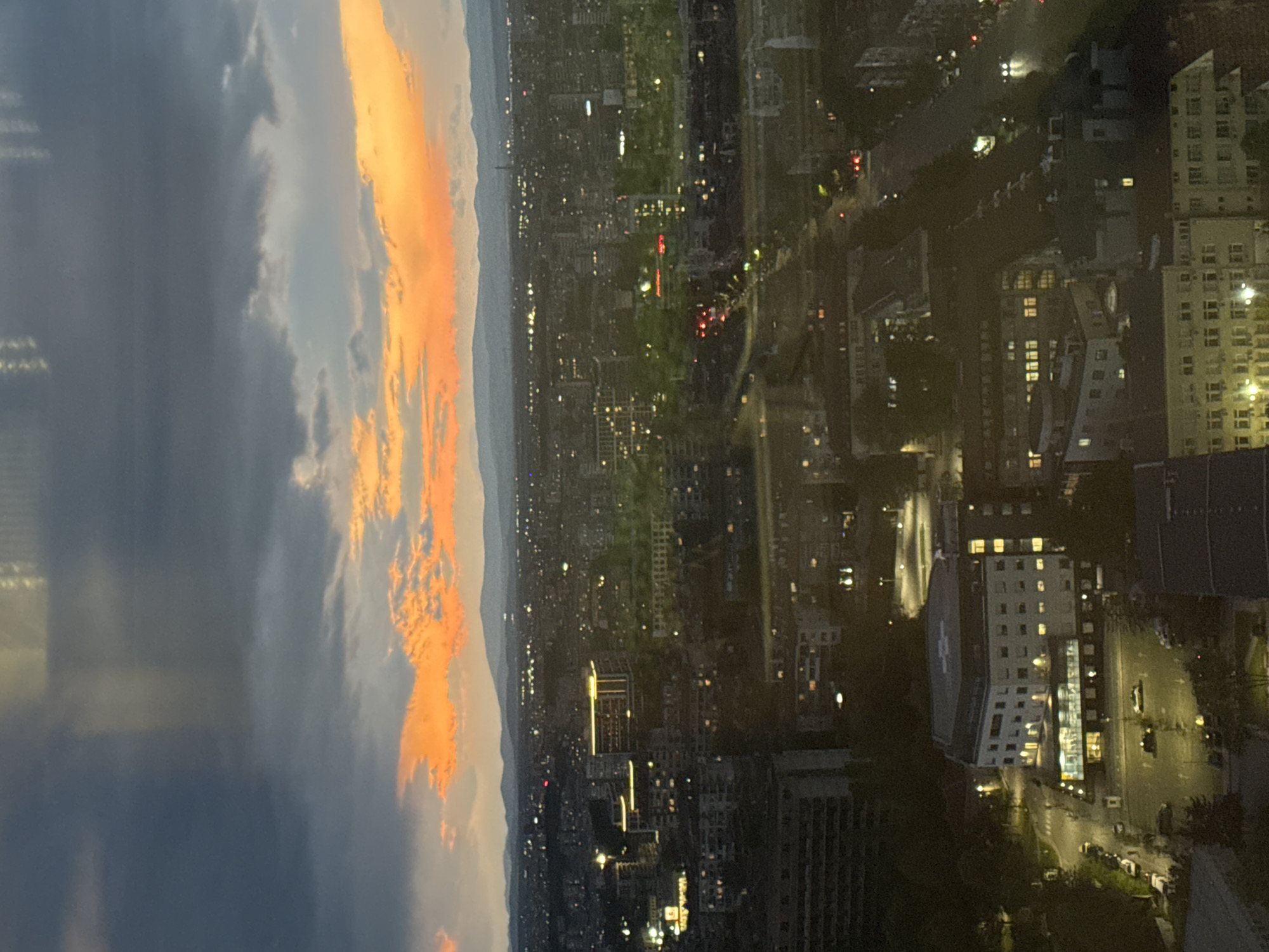 The image captures a cityscape at dusk, likely taken from a high vantage point in Sofia, Bulgaria. A dramatic sunset fills the left portion of the frame, with orange and yellow clouds transitioning to darker hues. The city below is a mix of residential and commercial buildings, interspersed with green spaces and streets. The lights of the buildings and vehicles create a network of illumination across the landscape. Near the bottom of the frame is what is likely a hospital due to the cross shape.
