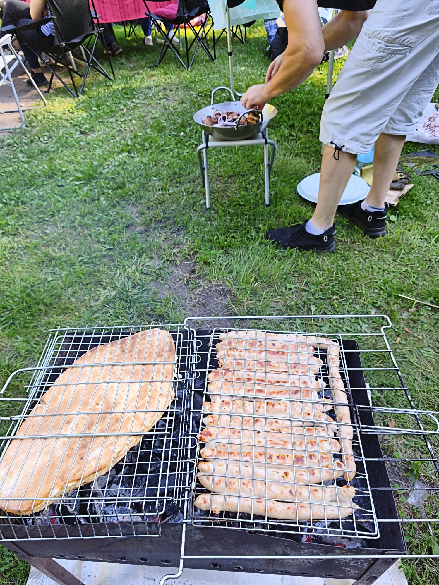 In Maardu, Estonia, an outdoor barbecue is in progress on a grassy area. A person in light-colored shorts and black sneakers is tending to food cooking in a metal pan on a small stand. In the foreground, a charcoal grill holds a large flatbread on the left side and numerous sausages arranged on a grill rack on the right side. The sausages are beginning to brown, indicating they are cooking. The surrounding environment is green grass, with several folding chairs visible in the background, suggesting a gathering or picnic. The lighting indicates it is daytime.