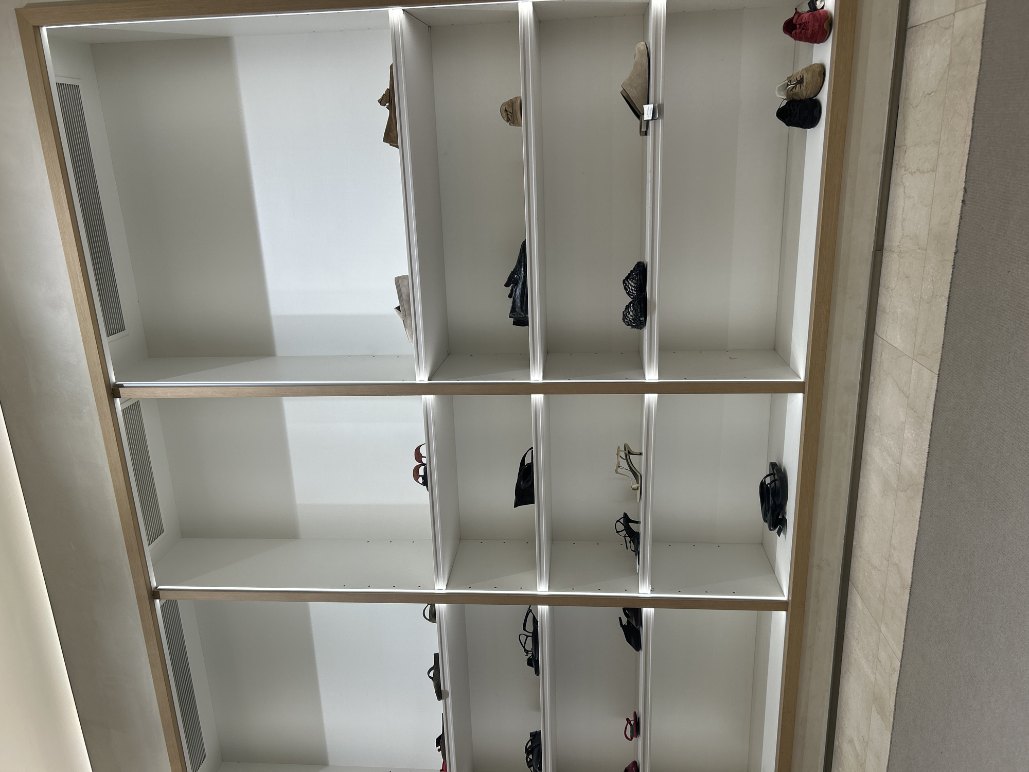 The image shows a built-in shoe rack with multiple shelves and compartments. The rack is primarily white, with a wooden trim around the edges of the shelves and vertical dividers. The shelves are occupied by an assortment of shoes, including sneakers, sandals, heels, and loafers, arranged somewhat haphazardly in the compartments. The location is not identifiable from the image.