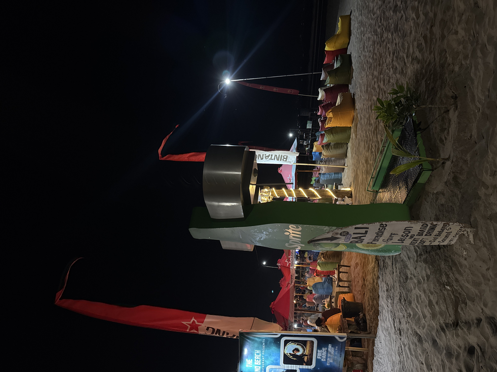 The image depicts a nighttime scene on a sandy beach in Kuta, Indonesia. In the foreground, a large, green, bottle-shaped sign with the word "Sprite" is prominently displayed. Adjacent to it, a weathered white sign lists services such as "BALI Paradise Surf Lesson", "RENT BOARD", "SOFT DRINK", "Cool Bintang", and "Coconut". A collection of colorful beanbag chairs are arranged on the sand, facing a row of red umbrellas providing shade. Behind the beanbag chairs, tables and people can be seen in the dim lighting, suggesting an outdoor dining or seating area. Two red flags with white designs are visible, one on the left and another further back, mounted on poles. A bright light illuminates the scene from a pole in the background, creating lens flares. The beach appears to be a commercial area with several businesses operating at night.