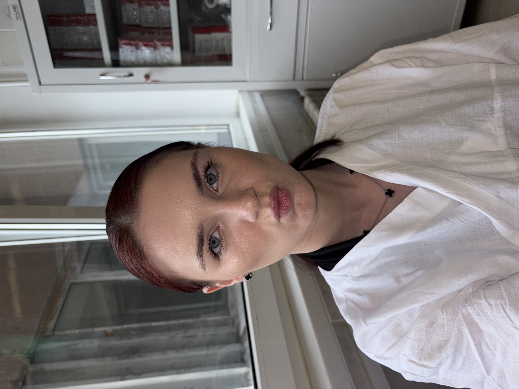 A medium shot captures a young woman with reddish-brown hair pulled back, posing for a selfie in what appears to be a medical or laboratory setting. She is wearing a white lab coat over a black undershirt and a necklace with a clover-shaped pendant. Her lips are pursed, and she is looking directly at the camera. The background shows a window with a metal structure outside, a white cabinet with glass doors displaying items, and a radiator. The overall lighting suggests an indoor environment.