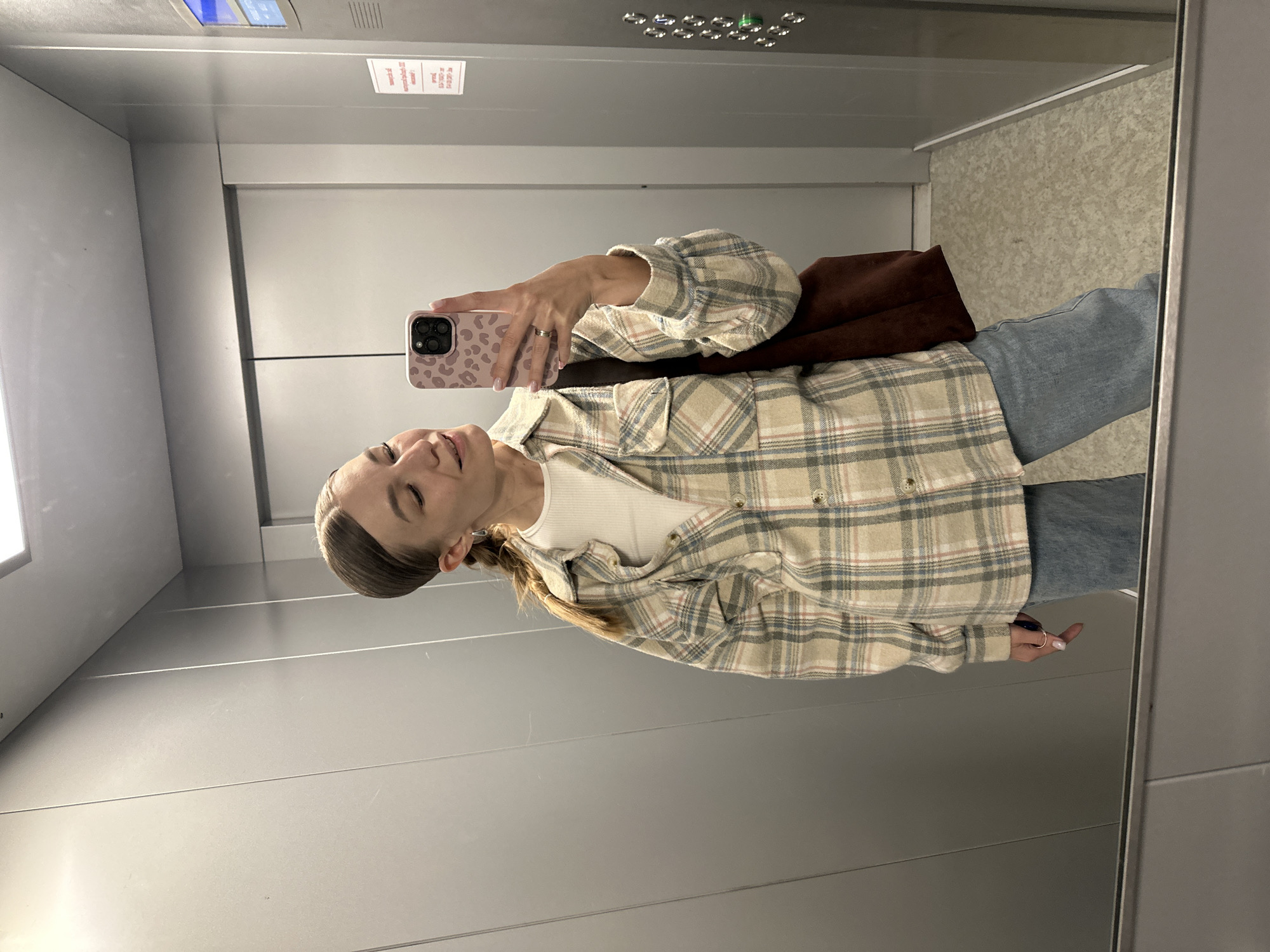 A young Caucasian woman with her hair pulled back in a braid stands in an elevator, taking a selfie in a mirror. She is wearing a cream-colored, long-sleeved plaid shirt over a white top, light blue jeans, and a brown tote bag is slung over her left shoulder. She is looking down at her phone, which has a pink leopard print case. The elevator walls are metallic gray, and the floor is a speckled light gray. The image appears to have been taken in an elevator in Chaikovsky, Russia.