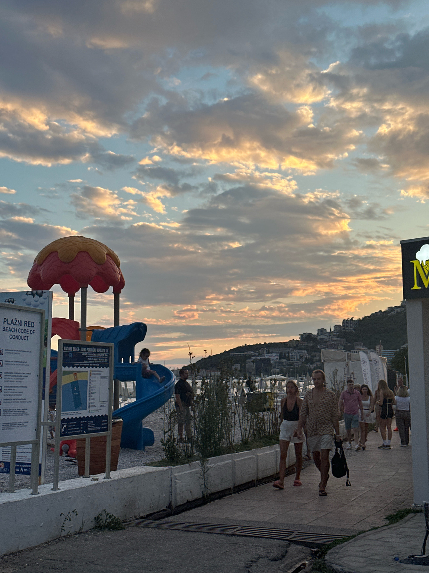 The image depicts a coastal scene in Budva, Montenegro, during what appears to be late afternoon or early evening. The sky is filled with dramatic, cloud-laden sunset colors, featuring hues of orange, yellow, and gray against a pale blue backdrop.
In the foreground, to the left, is a playground structure with a red and yellow mushroom-shaped canopy and a blue slide. A young child is visible on the slide, and a man stands to the right of the playground. Adjacent to the playground are several signs. One sign, predominantly white, has black text that appears to be a beach code of conduct, with the title "PLAŽNI RED BEACH CODE OF CONDUCT" in larger font. Another sign, blue and white, also contains text and a map of the beach.
To the right of the playground, a paved walkway runs along the coast. Several people are walking on this walkway. A couple in the center are prominent: a man wearing a patterned shirt and shorts, carrying a black bag, and a woman in a dark top and white shorts. Other individuals and groups are also walking along the path.
Beyond the walkway and playground, a body of water (likely the Adriatic Sea) is visible, with boats docked in a harbor. In the background, the hilly coastline of Budva is dotted with buildings. A bright sign with the letter "M" and a cloud-like symbol is visible on the far right of the image, partially obscuring the buildings. The overall environment suggests a popular tourist area with recreational facilities and coastal amenities.