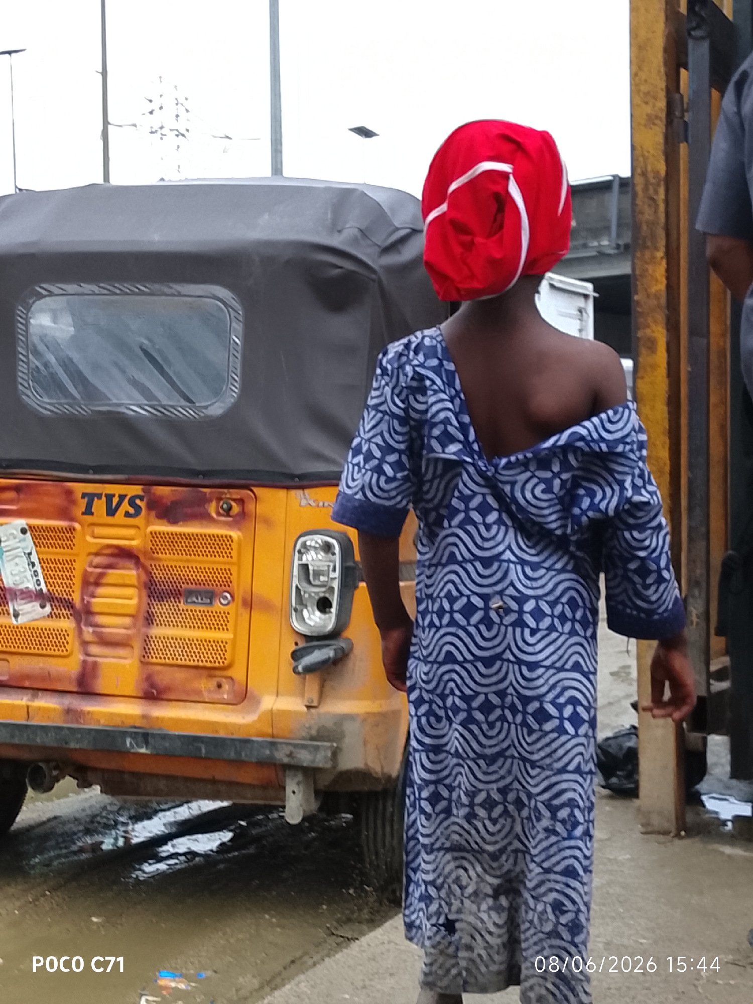 In Lekki, Nigeria, an individual from the rear wears a vibrantly colored red headwrap adorned with white trim, and an off-the-shoulder blue and white patterned dress. They stand beside a yellow auto-rickshaw, which features the "TVS" logo. The auto-rickshaw shows signs of wear and tear, with a distressed texture and some smudges. The environment appears to be an urban setting with a paved surface, partially covered in muddy water, indicating recent rainfall or a drainage issue. In the background, a metal structure, possibly a gate or fence, stands behind the individual. The overall scene is captured outdoors, suggesting a street-level perspective.