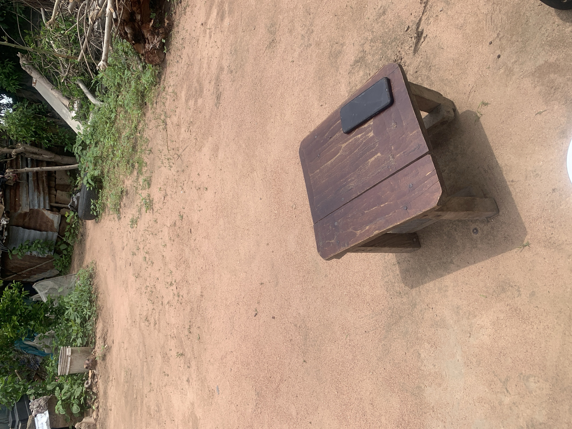 The image depicts an outdoor scene on a dirt path in Jalingo, Nigeria. In the foreground, a dark wooden stool with visible wood grain and nail heads sits on the sandy ground. A black smartphone is placed on the surface of the stool. The ground is a light brown, dusty earth with scattered small green weeds. In the background, the scene is cluttered with various items including corrugated metal sheeting, green vegetation, a bucket, tires, and a pile of logs and branches. The overall impression is one of an outdoor, possibly rural or informal setting, with remnants of construction or discarded materials visible. The lighting suggests it is daytime, with shadows cast by the stool and objects in the background.