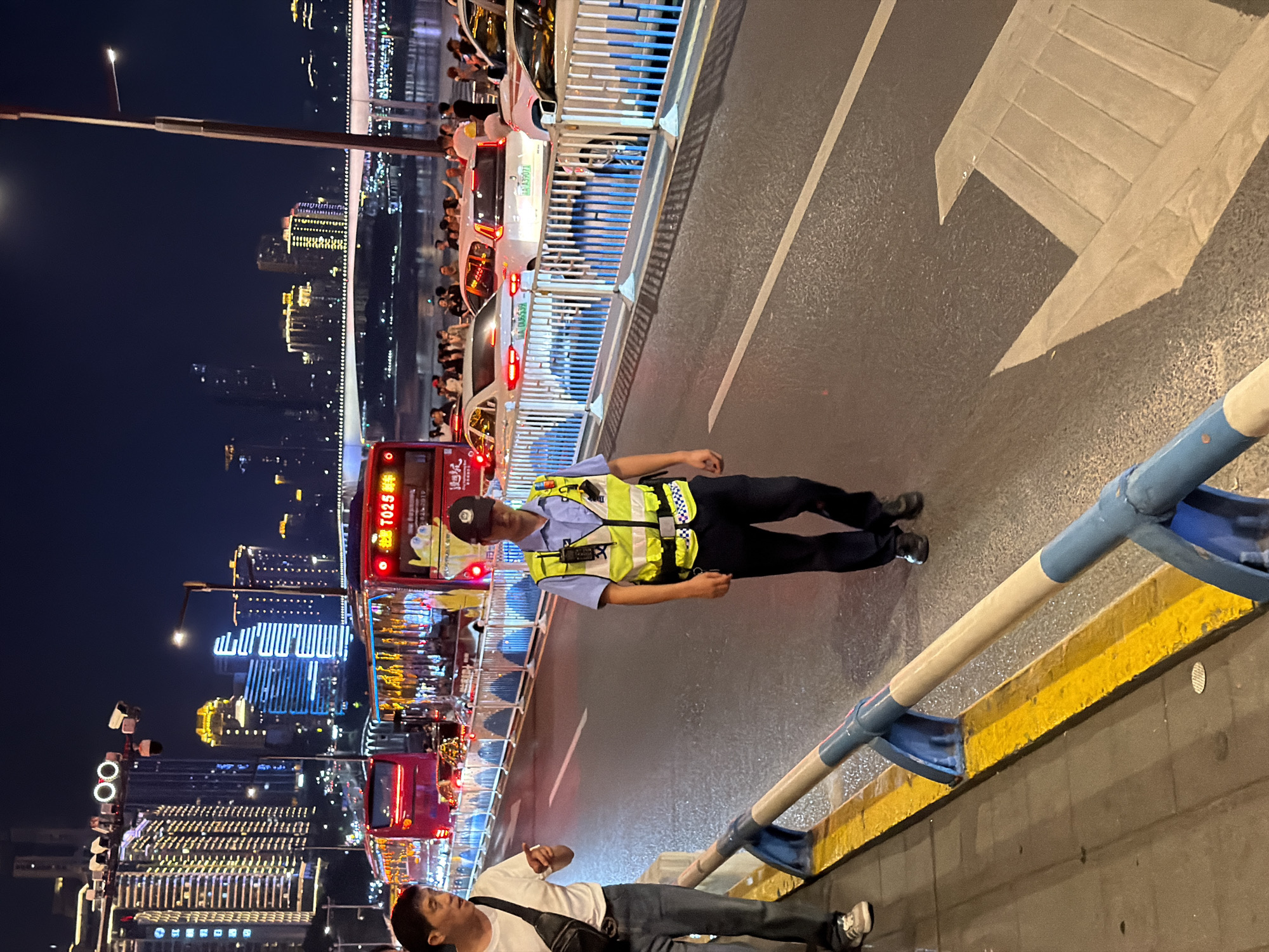 This image captures a nighttime scene on a busy road in Yuzhong District, China, likely a bridge or elevated roadway given the cityscape and river visible in the background. In the foreground, a police officer wearing a bright yellow safety vest over a blue uniform walks towards the right of the frame. He is wearing a dark cap and dark pants. To his left, a man in a white t-shirt and jeans, with a black crossbody bag, walks away from the camera, holding a cigarette.
The road is wet, indicating recent rain or cleaning. Several vehicles, including red buses and white cars, are visible on the road, suggesting traffic. A barrier with blue and white vertical posts separates the pedestrian walkway from the road. In the background, illuminated skyscrapers form a dense urban skyline, reflecting light onto the scene. The overall atmosphere is one of urban activity at night, with the presence of law enforcement and general public.