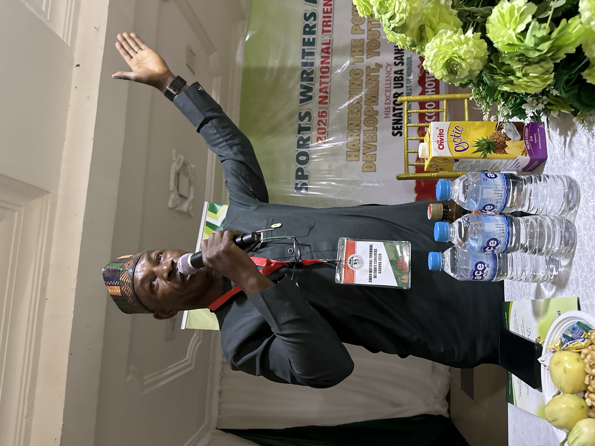 A man wearing a dark green traditional attire and a patterned cap stands on a raised platform, speaking into a microphone. He has his right arm raised, with his hand open and palm facing outward, as if gesturing or emphasizing a point. A lanyard with a badge is around his neck. In front of him, on a table covered with a white tablecloth, are two bottles of "Ace" water, a carton of "Chivita Exotic" pineapple and coconut juice, a smartphone, and a plate containing some light-colored fruits and nuts. Behind the man, a banner displays text related to "SPORTS WRITERS" and a "2026 NATIONAL TRIENNIAL" event, with a subtitle about "HARNESSING THE POWER OF..." and mentioning "HIS EXCELLENCY SENATOR UBA SANI". The background appears to be an indoor venue with white walls and ceiling. Green drapes are visible on the left side of the image. The lighting is artificial, suggesting an indoor event. The overall scene depicts a formal presentation or speech at an event in Barden Yana, Nigeria, given the mention of Kaduna and Senator Uba Sani, who is the governor of Kaduna State.