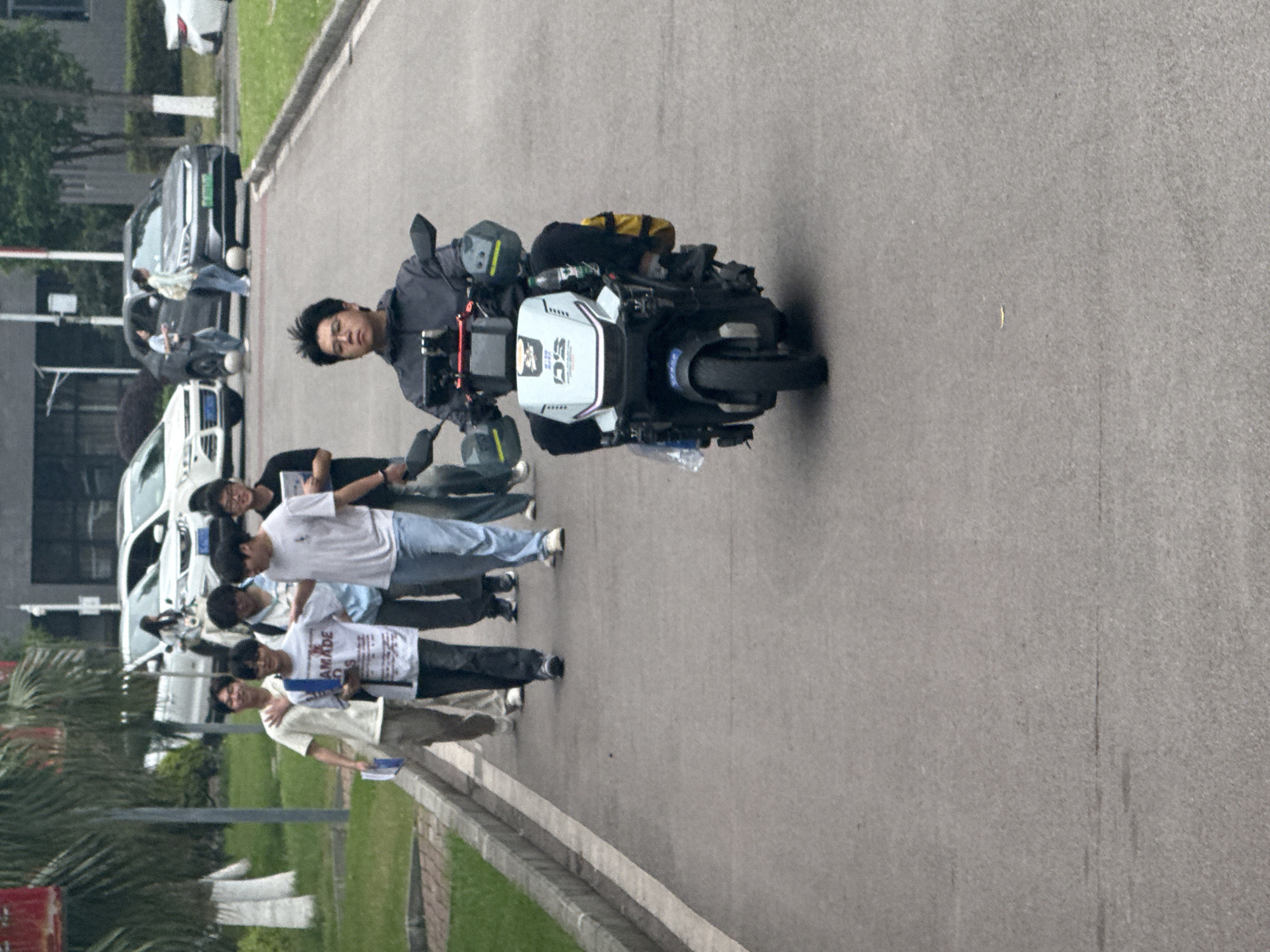 The image shows an Asian man riding an electric unicycle on an asphalt road in Jiangjin, China. The rider is looking at the camera, and the unicycle is equipped with luggage and equipment. A group of young men are walking along the side of the road. There are buildings, cars, and greenery in the background.