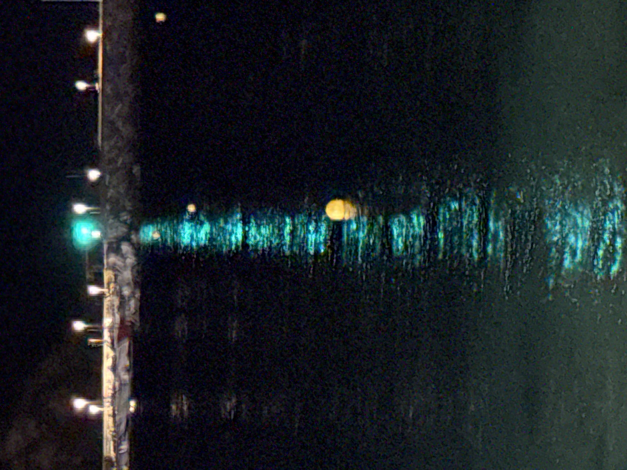 At night in Blanes, Spain, a body of water reflects the illuminated structures and lights of a pier or seawall. Multiple bright, white lights are visible along the elevated structure, some appearing as single points and others as clusters. A few yellow buoys or floating objects are present in the water, with one prominent yellow sphere reflecting brightly. The water itself exhibits a strong turquoise luminescence, suggesting the presence of underwater lighting or reflections from the structures above. The overall scene is characterized by darkness punctuated by artificial light sources and their reflections on the calm water.