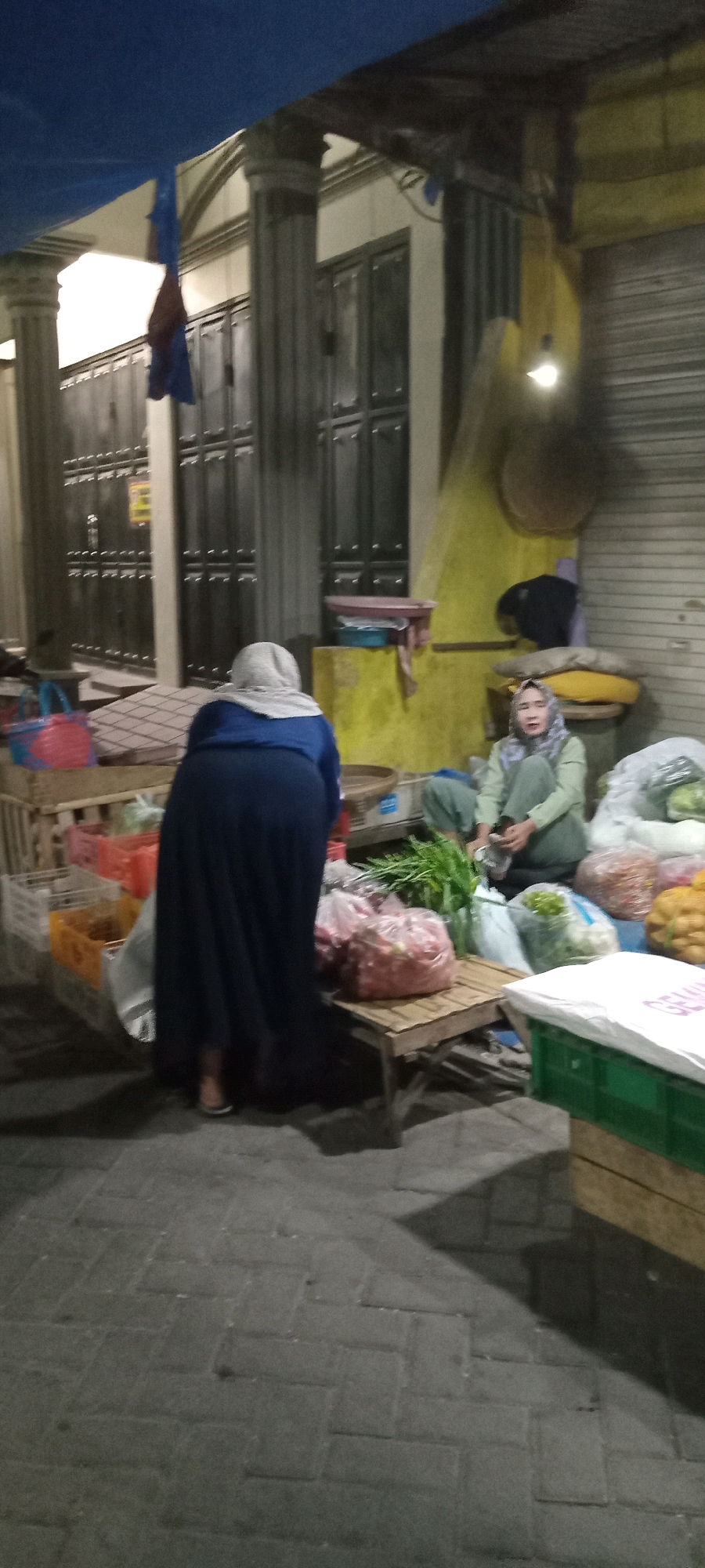The image depicts a nighttime scene at a market stall in Babat, Indonesia. In the foreground, a person wearing a blue long-sleeved shirt and a dark blue skirt, with a light grey hijab, is bent over, facing away from the camera. They appear to be interacting with the items on the stall. To the right, another person, wearing a light green long-sleeved shirt and a grey hijab, is seated and appears to be bagging produce. The stall itself is laden with various goods, including bags of what appear to be onions, leafy green vegetables, and other produce. Red and blue plastic baskets are visible on the left side of the stall. The ground is paved with rectangular grey bricks. In the background, a building with large doors and pillars is visible, along with a corrugated metal shutter. A blue tarp is stretched overhead. The lighting suggests artificial light sources, casting shadows across the scene.