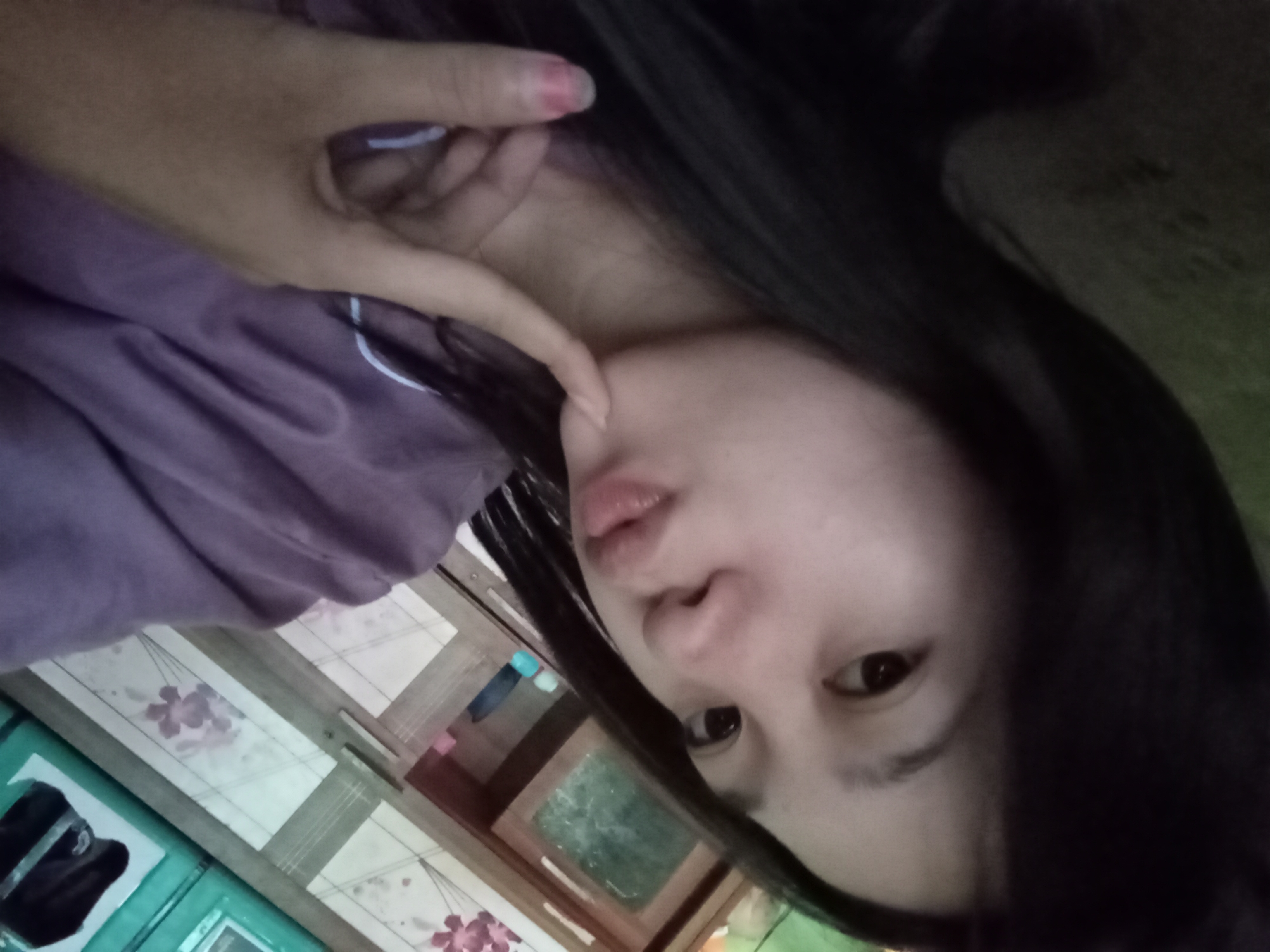 A young Indonesian woman is taking a selfie. She has long, dark hair and is wearing a purple pajama top. Her right hand is resting on her chin, and her left hand is near her face. The background appears to be a room in a house in Rejosari, Indonesia, with wooden furniture and floral decorations visible.