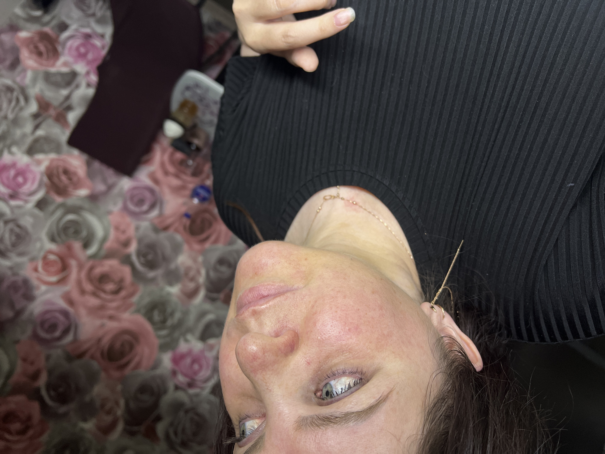 A close-up, low-angle shot features a woman with fair skin and gray-blue eyes, gazing upwards. Her face exhibits some redness, particularly around her nose. She is wearing a black ribbed top and a delicate gold necklace. A long, thin gold earring dangles from her left ear. The background is a floral pattern with roses in shades of pink, purple, and gray. The location is Noviy, Russia.