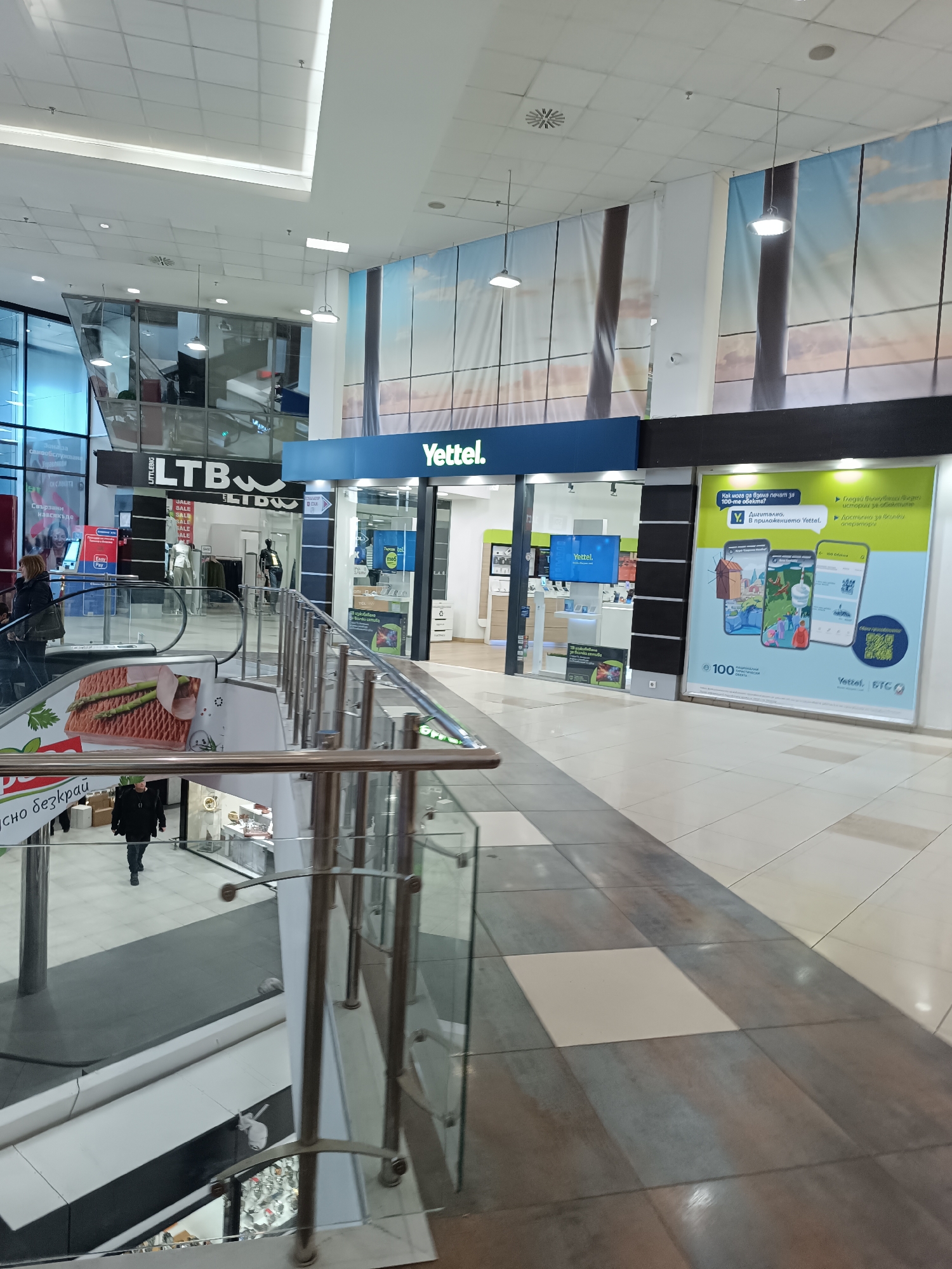 The photograph captures an interior view of a shopping center in Sofia, Bulgaria. The perspective is from a second-level walkway overlooking the first level. The Yettel mobile store is visible in the foreground, located beside an LTB clothing store, both of which are accessible from the walkway. The stores have storefronts with signage in Cyrillic.
The Yettel store displays promotional material for its services. An escalator is to the left of the frame, and a poster of a ham sandwich is adhered to its side. A man is walking on the level below, passing by a jewelry store, which is visible through a glass railing on the walkway. The flooring of the walkway is tiled in a alternating squares. The ceiling of the shopping center is high, with linear lighting fixtures and decorative landscape banners.