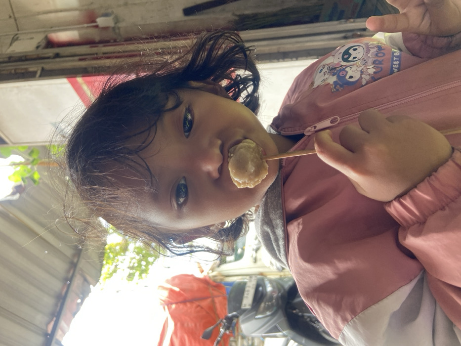 A young girl with dark hair and eyes holds a meatball on a stick to her mouth. She wears a pink jacket with cartoon characters and white panels. The background features a canopy, trees, and a blurred scooter or motorcycle. The image has a slightly tilted angle and bright lighting.