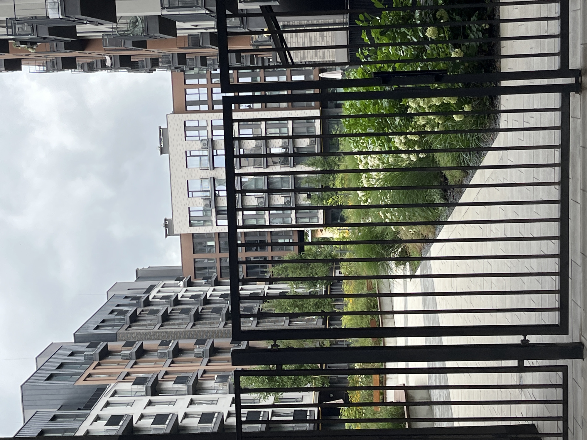 A black metal gate in the foreground of the image opens onto a courtyard of modern apartment buildings in Lviv, Ukraine. The buildings are multi-story with balconies and large windows, constructed with a mix of light-colored and dark brick facades, with some sections featuring copper-toned metal cladding. The courtyard itself features paved pathways, lush green landscaping with tall grasses and flowering bushes, and some trees. The sky is overcast and gray. The image appears to be taken during daylight hours.