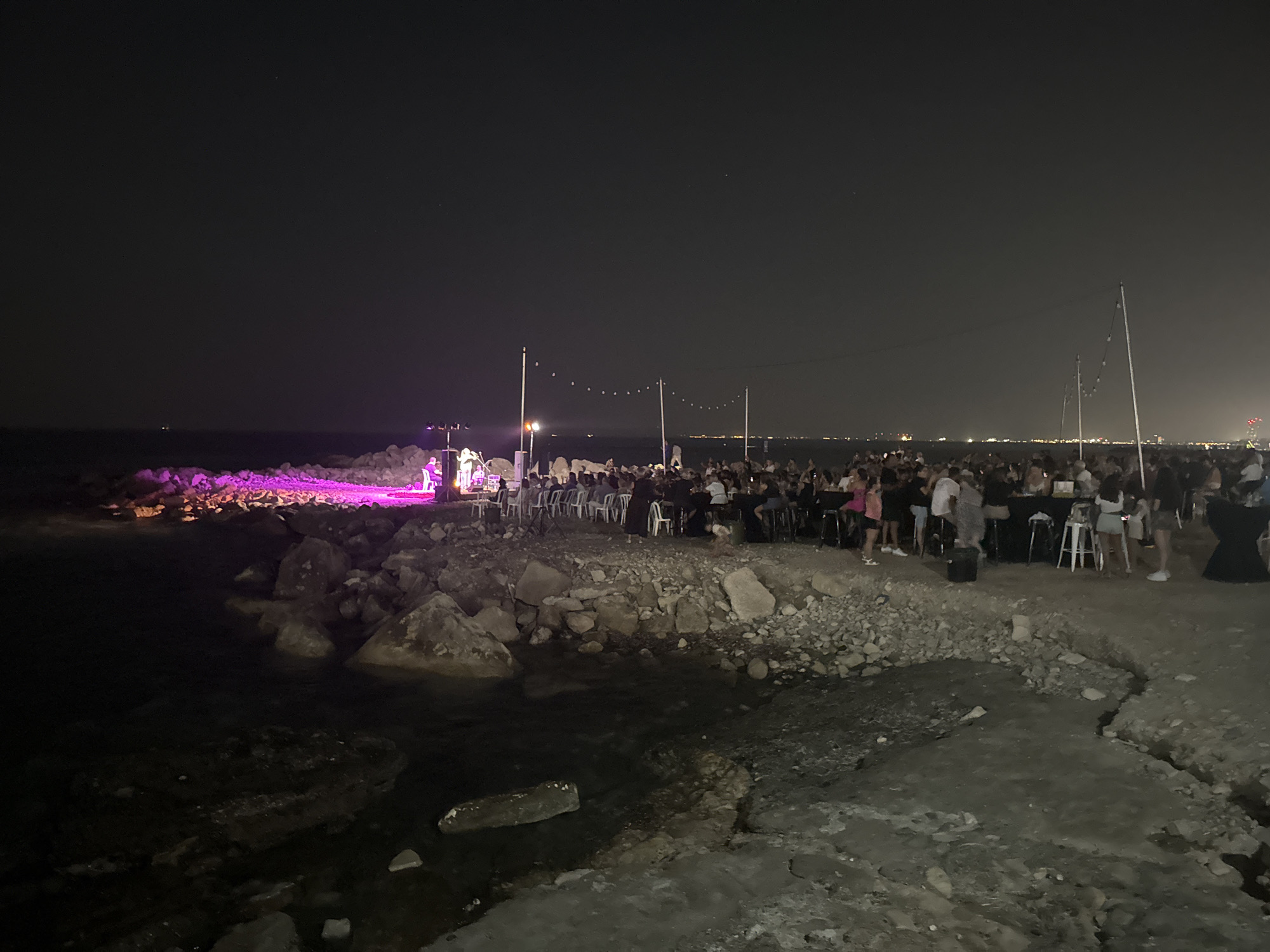 An outdoor event is taking place at night on a rocky shoreline in Agios Tychona Community, Cyprus. A stage with bright pink lighting is set up on the left side of the image, where a band appears to be performing. A crowd of people is gathered in front of the stage and further along the shoreline, seated at tables and standing. String lights are suspended in the air above the seating area. The sea is visible on the left, with calm water lapping against the rocks. The overall atmosphere suggests a lively gathering, possibly a concert or a party, held by the water under the night sky.