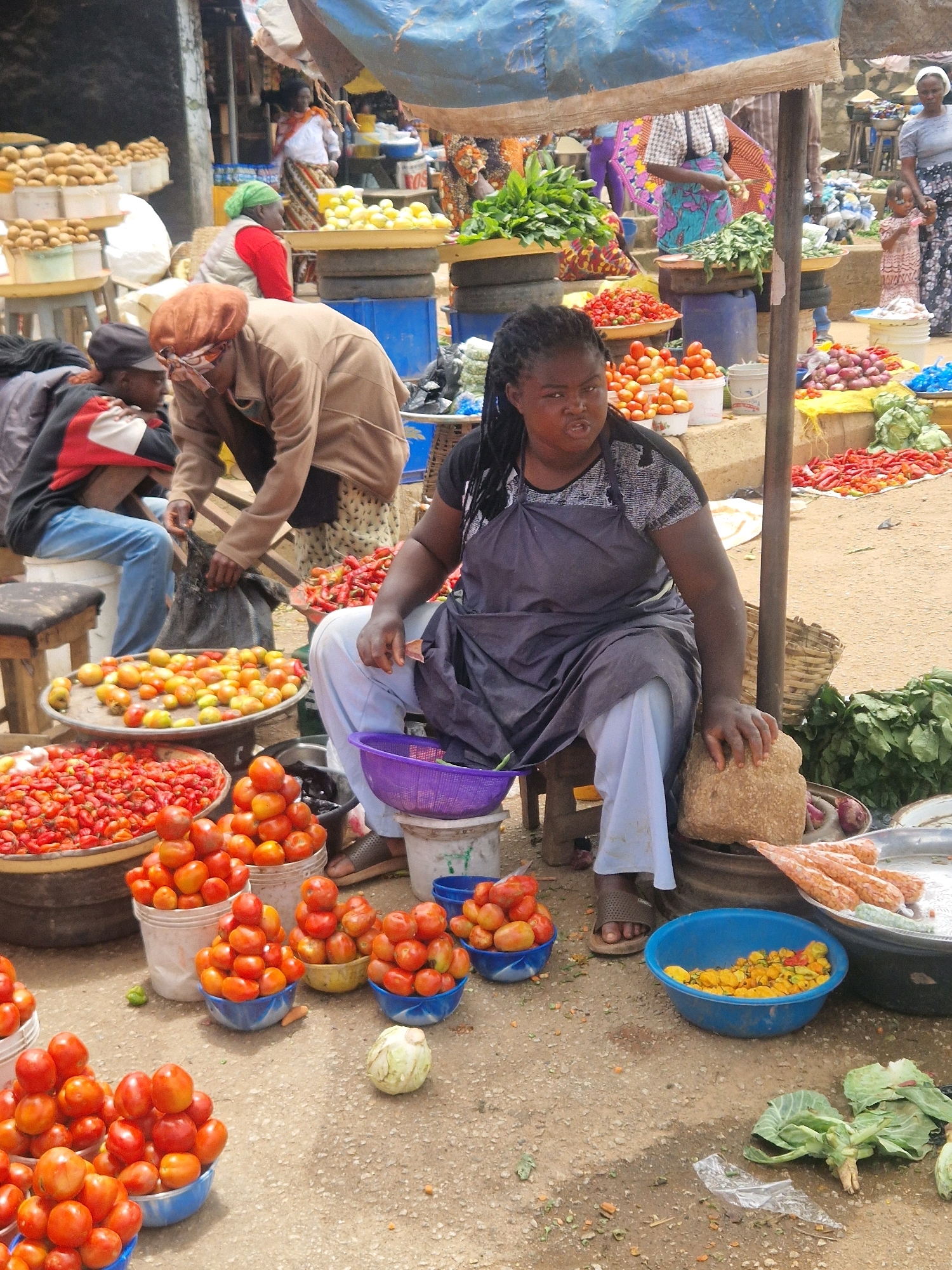 In an outdoor market scene, a woman with dark skin and braided hair, wearing a dark gray apron over a patterned shirt and light blue pants, sits on a stool in the foreground. She is engaged in grinding something with a large stone on top of a dark metal drum. Beside her, to the left, are two buckets and a purple bowl filled with tomatoes and peppers. In front of her, to the right, a blue bowl contains small yellow peppers, and a large metal tray holds what appear to be pinkish sausages or processed meat. The ground is dirt and gravel, littered with vegetable scraps and plastic. Behind the woman, to the left, are other market stalls with various produce, including piles of tomatoes, red peppers, and onions. Several people are visible in the background, some appearing to be vendors and customers. The market is bustling with activity, indicated by the assortment of goods and the presence of multiple individuals.