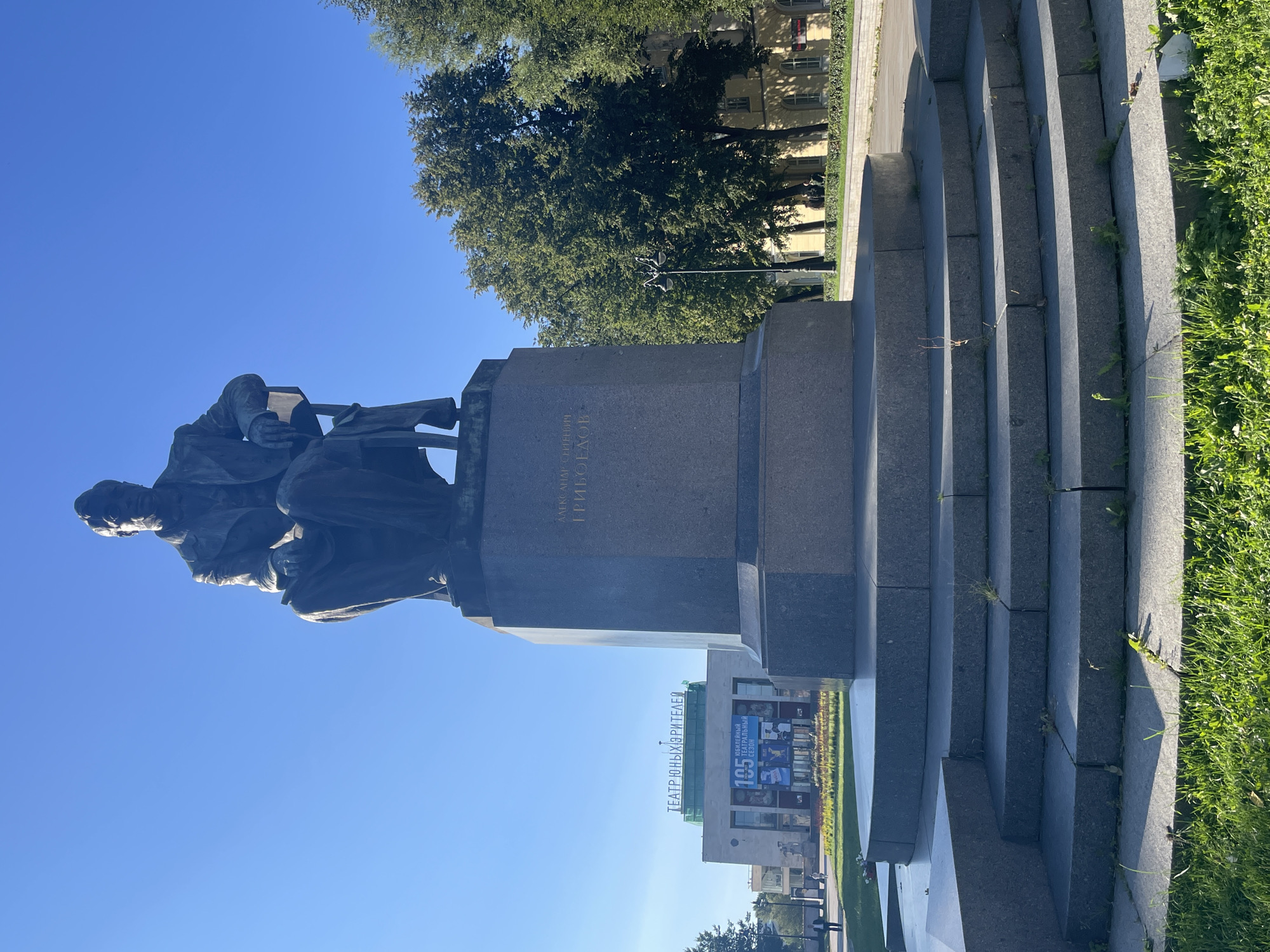 The image depicts a bronze statue of Alexander Griboyedov, a prominent Russian playwright, poet, and diplomat, seated on a chair and holding a book. The statue is situated in front of a large, modern building in Saint Petersburg, Russia, identified by the Cyrillic inscription "ТЕАТР ЮНЫХ ЗРИТЕЛЕЙ" (Theater of Young Spectators) above its entrance. The building features a large banner with the number "105" and Cyrillic text, indicating a significant anniversary related to the theater. The statue stands on a granite pedestal, accessed by a series of stone steps. Lush green grass and trees surround the monument and the building. The sky is clear and blue. The overall scene appears to be a public square or park area within the city.