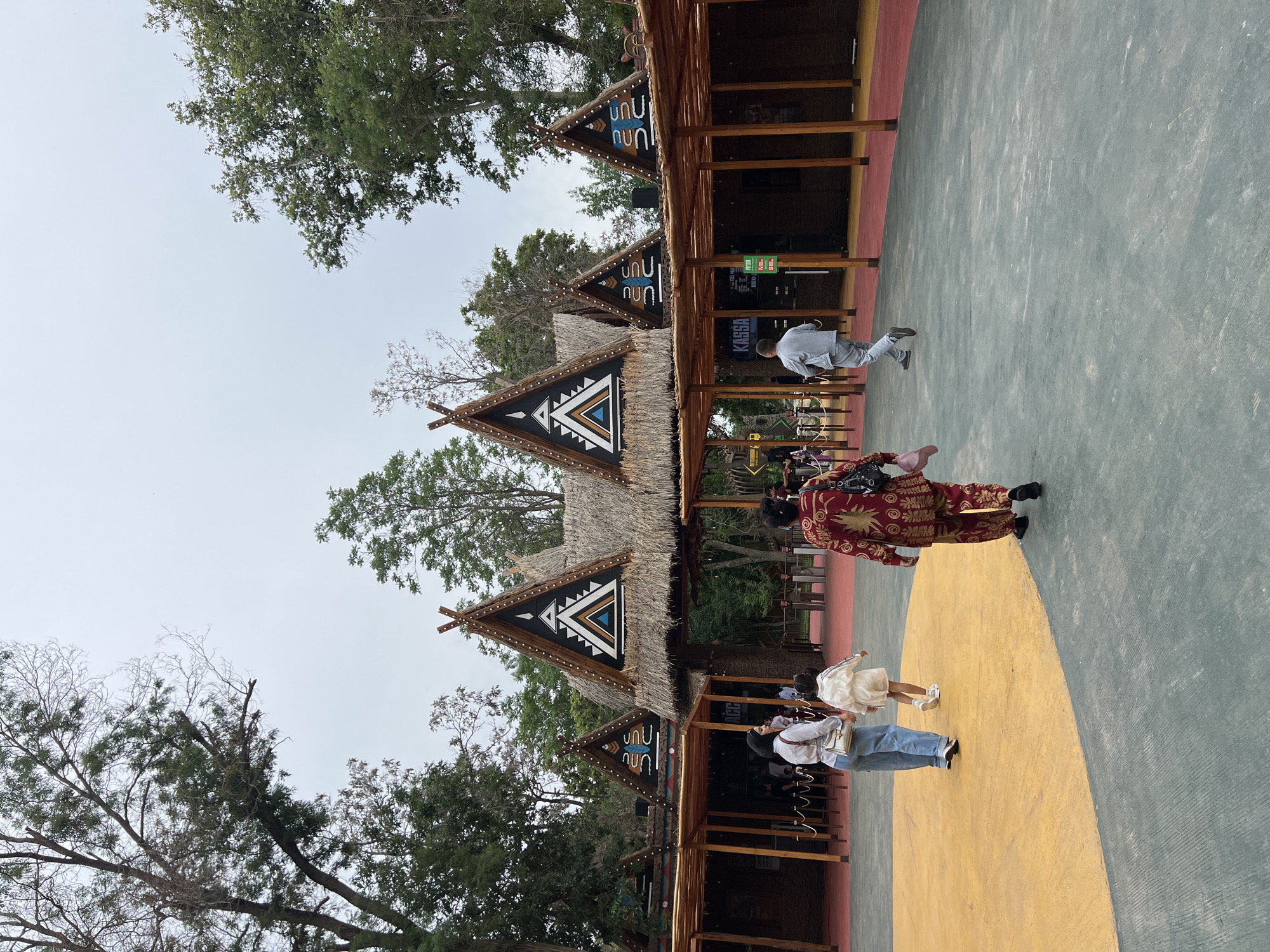 The image shows an outdoor scene in Tashkent, Uzbekistan, likely within a park or tourist attraction featuring structures with thatched roofs. The architecture suggests an ethnic or cultural theme.
People: Several individuals are present. A person in a red patterned garment is walking towards the right. A woman is seen interacting with a young child, who is walking on a yellow surface that seems to be painted on the ground. Another person wearing grey clothes walks away from the camera at a faster pace.
Objects: There are stalls, each featuring triangular thatched roofs with intricate tribal-like geometric designs. A ‘KASSA’ sign is visible in German. Signs listing prices are also visible. Trees are present in the background, adding to the natural setting.
Environment: The ground is divided into color-blocked sections of yellow, grey, and red. The sky is overcast. The presence of trees suggests a park-like or garden-like area.