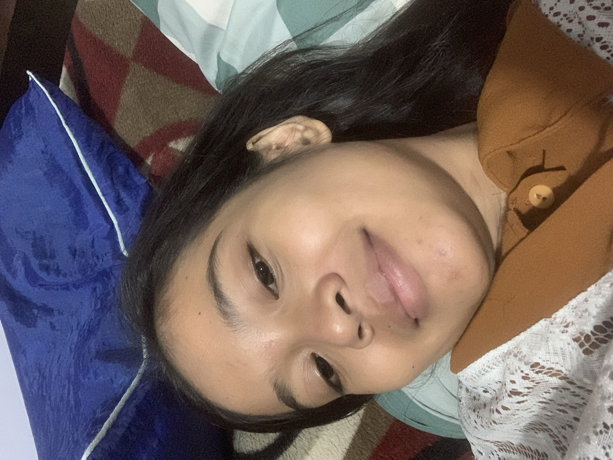 A young woman with dark hair and brown eyes lies on her back, looking at the camera with a slight smile. She is wearing a white lace top and a rust-colored button-up shirt. The background consists of a blue pillow and a patterned blanket. The lighting suggests an indoor setting, possibly a bedroom. The image was captured in Sungai Meriam, Indonesia.