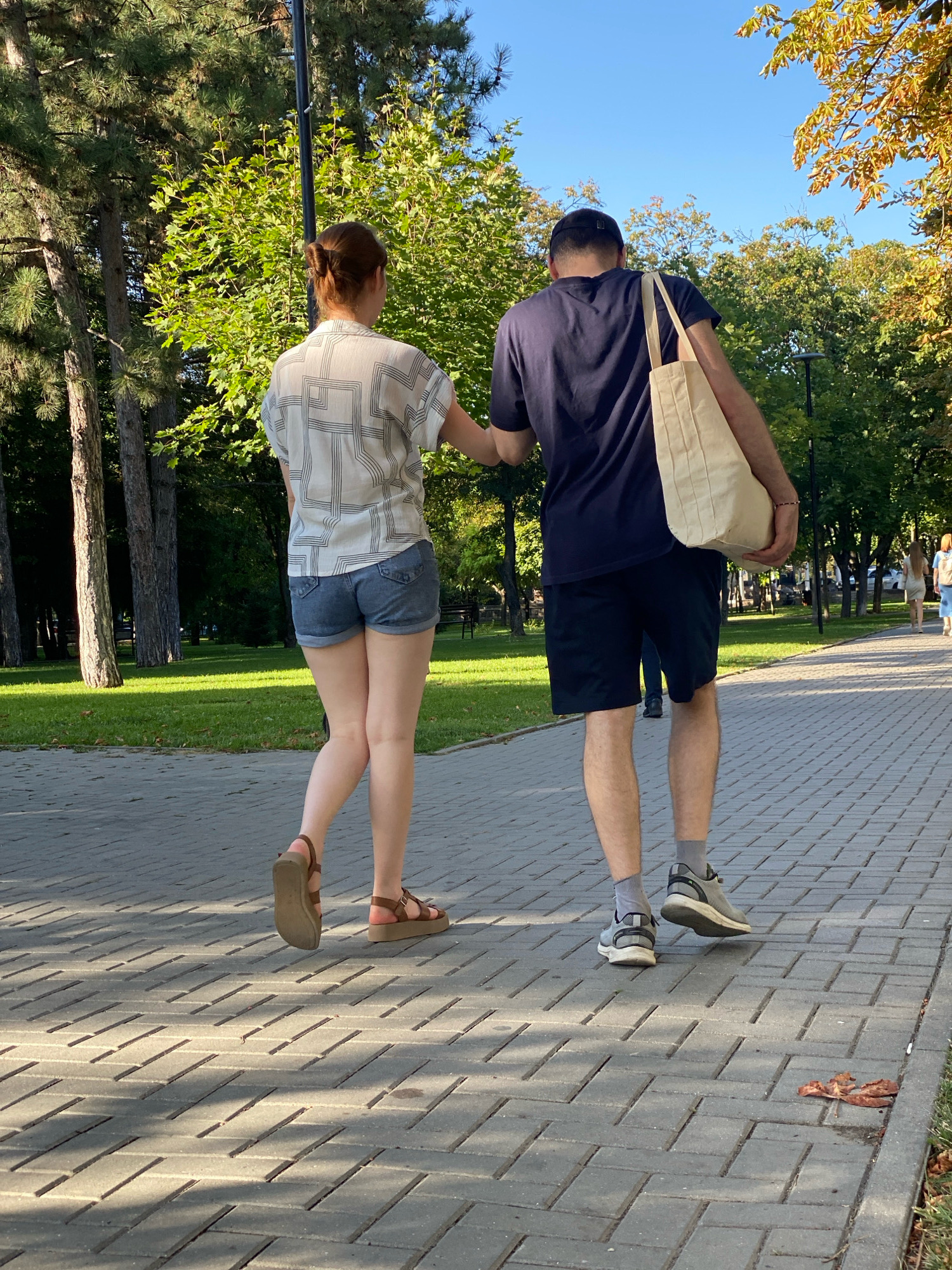Two individuals, a woman and a man, are walking on a paved pathway in a park. The woman, facing away from the camera, has her hair in a bun and is wearing a white patterned short-sleeved shirt and denim shorts. She is wearing brown sandals. The man, also facing away, is wearing a dark blue t-shirt, dark shorts, and gray athletic shoes with gray socks. He is carrying a light-colored tote bag over his right shoulder. They are walking side-by-side, with the woman's right arm lightly touching the man's left arm. The pathway is made of gray interlocking pavers. The background shows a lush green park with mature trees, some with yellowing leaves, suggesting it is autumn. The sky is clear and blue. The scene appears to be set in a public park in Chișinău, Moldova, given the common architectural and landscape features associated with the city.