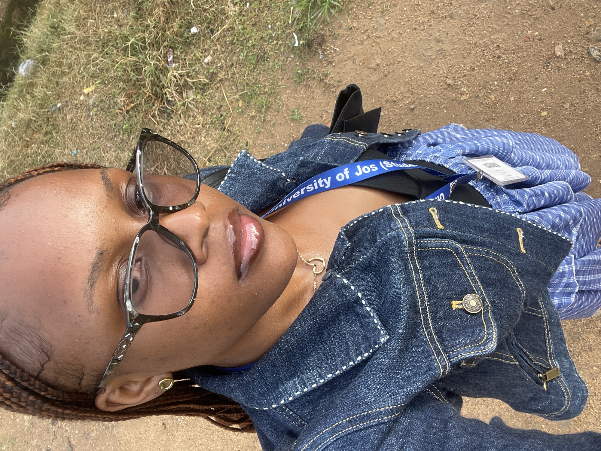 A young Black woman with braided hair and glasses stands outdoors on a dirt path with some dry grass and debris in the background. She is wearing a denim jacket over a black top and a blue and white plaid skirt. A blue lanyard with "University of Jos (Stud..." printed on it is around her neck, and a white ID card with a QR code hangs from it. She is looking towards the camera with a slight smile. The setting appears to be a rural or semi-urban area, potentially on the outskirts of the city of Rafin Par, Nigeria.