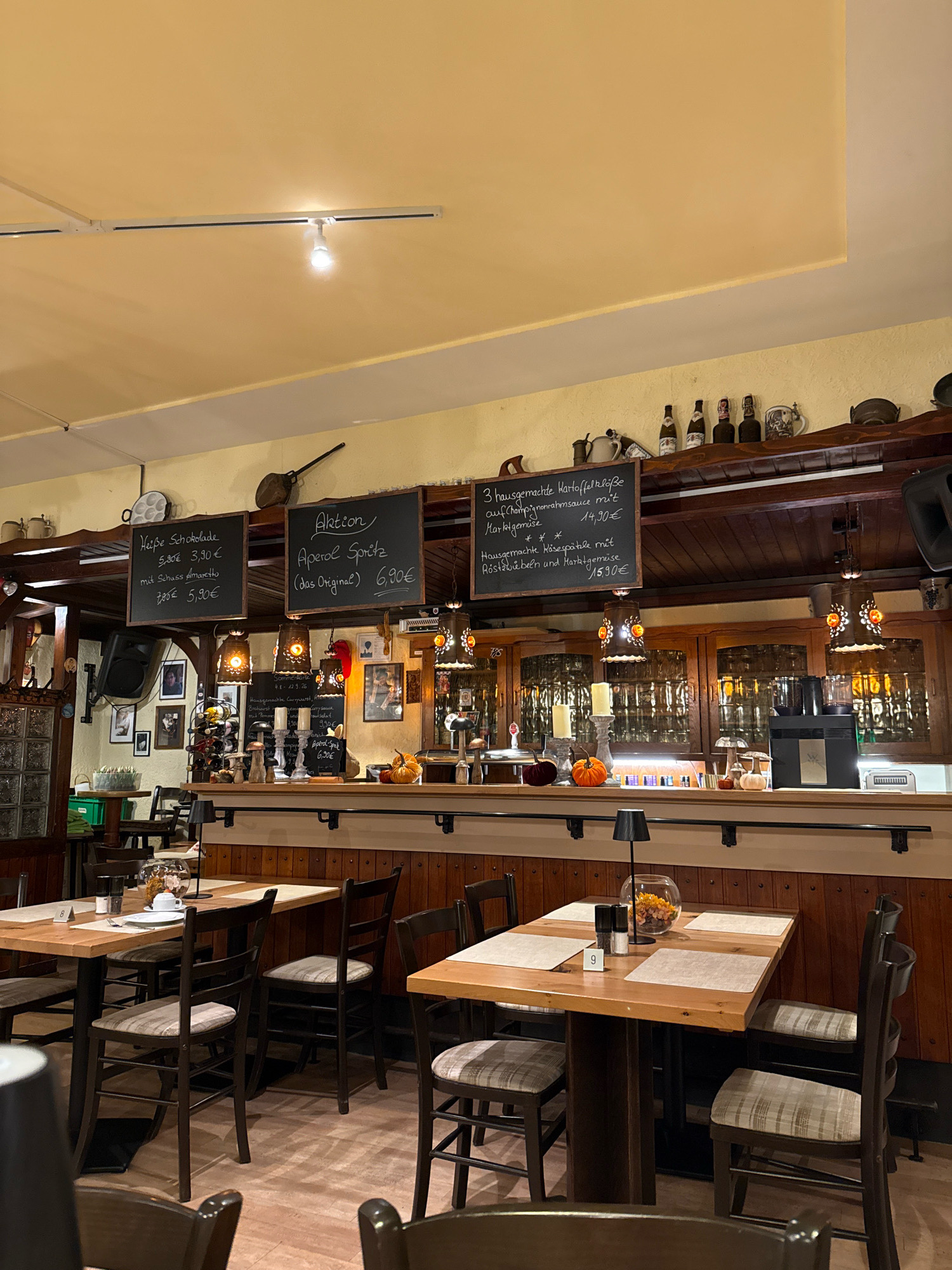 The image depicts the interior of a restaurant in Bonn, Germany, specifically an establishment that appears to serve German cuisine and drinks. In the foreground, several wooden tables and chairs are set, suggesting a dining area. The tables are made of light-colored wood with placemats and salt and pepper shakers. One table has a small glass bowl filled with decorative autumn-themed items, including mini pumpkins.
In the background, a bar area is visible with wooden paneling and a wooden counter. Various bottles of beverages and decorative items are displayed on shelves above the bar. There are chalkboards hanging from the ceiling, displaying menu items and prices in German. One chalkboard advertises "Aktion Aperol Spritz (das Original) 6,90€". Another lists "Heiße Schokolade 5,50€ 3,90€" and "mit Schuss Amaretto 7,00€ 5,90€". A third chalkboard lists two dishes: "3 hausgemachte Kartoffelklöße auf Champignonrahmsauce mit Marktgemüse 14,90€" and "Hausgemachte Käsespätzle mit Röstzwiebeln und Marktgemüse 15,90€".
Several decorative lamps with intricate metalwork and colored glass hang from the ceiling, casting a warm light. Artwork and framed photographs are displayed on the walls. The overall ambiance is cozy and rustic, typical of a German beer garden or traditional restaurant. The lighting suggests it is evening or a dimly lit interior. There are no people clearly visible in the image, but the setup indicates a place prepared for customers. The flooring appears to be light-colored wood.