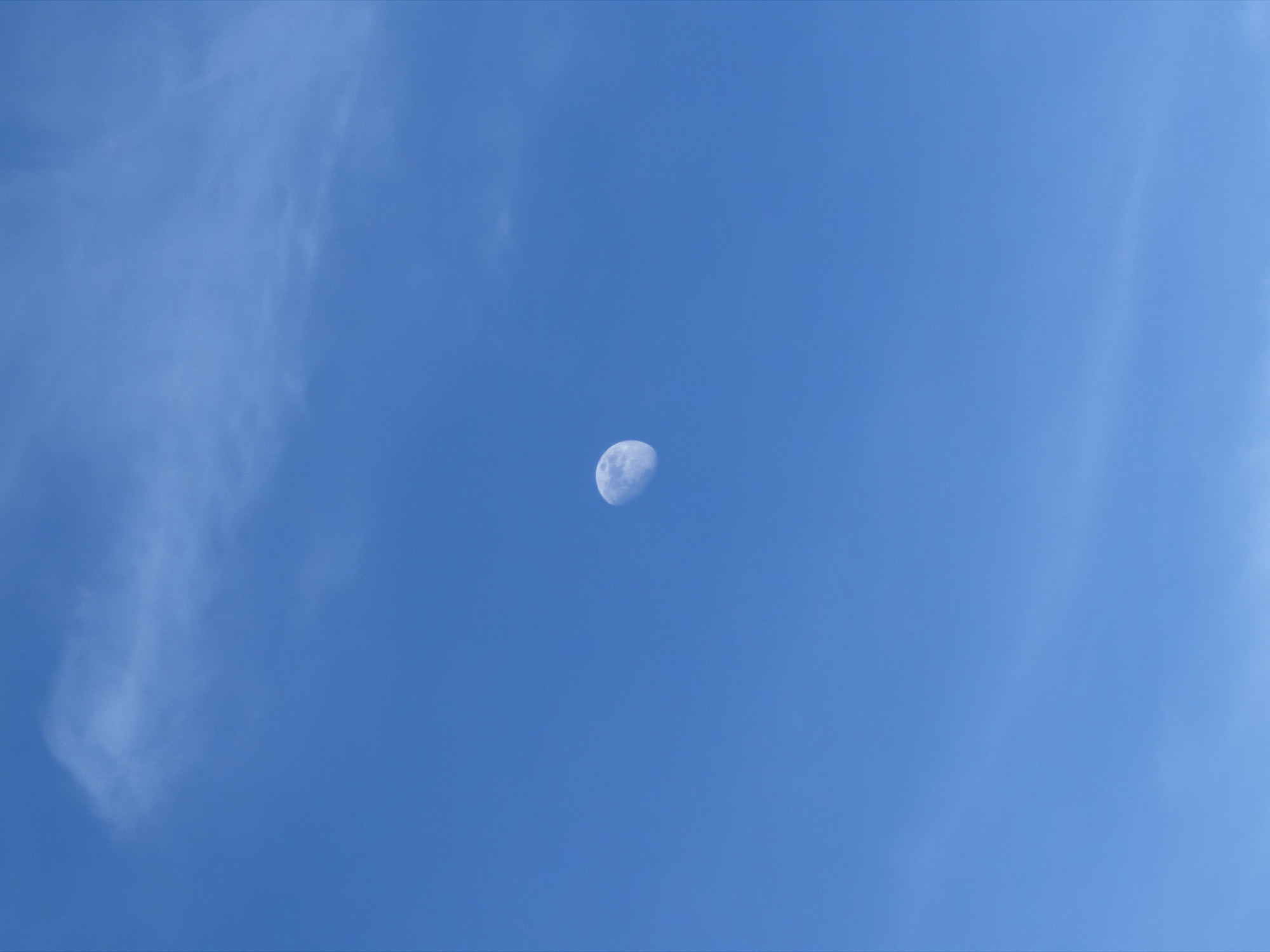 The image displays a daytime sky, presumed to be observed from Izhevsk, Russia, though no terrestrial landmarks are present to confirm a specific location within the city. The primary subject is the Moon, visible in a gibbous phase, positioned slightly above the central horizontal axis and to the left of the central vertical axis. The Moon appears off-white with visible crater features, illuminated against the bright blue sky. The sky itself is a clear, vibrant blue, traversed by thin, wispy cirrus-type clouds. A noticeable cloud formation extends from the upper left quadrant, and fainter, elongated cloud streaks are present in the mid-right and lower portions of the frame. There are no people, man-made objects, actions, or interactions depicted in the scene, which is solely focused on the celestial and atmospheric environment.