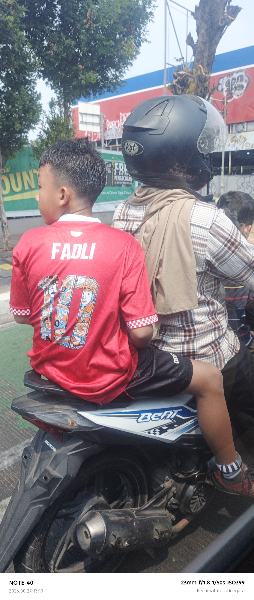 Two individuals are seated on a Honda Beat motorcycle, which is stationary on a paved road, possibly in a city environment. The person in the foreground is a young male wearing a red t-shirt with the name "FADLI" and the number "10" on the back. He also wears black athletic shorts. The person behind him is wearing a plaid long-sleeved shirt and a light brown scarf draped over their shoulders, along with a black helmet. The background includes trees, a building with a red and blue facade, and a street sign. The image was captured in Kecamatan Jatinegara, East Jakarta, Indonesia.