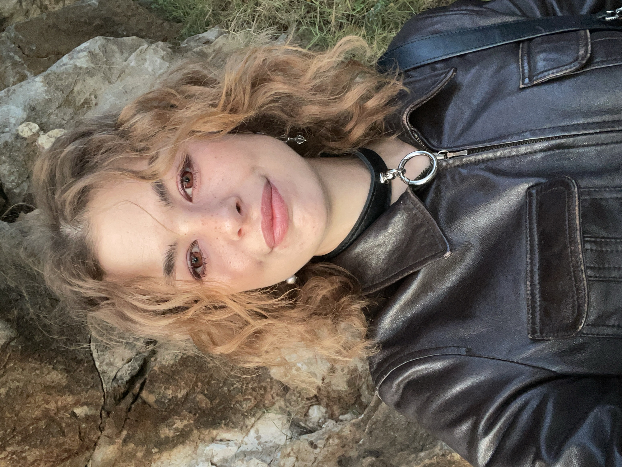 A young woman is depicted in a rotated close-up portrait, appearing to lie on her side outdoors. She has shoulder-length, wavy or curly hair, a mix of blonde and light brown tones. Her fair skin is adorned with noticeable freckles across her nose and cheeks. Her brown eyes, which exhibit a reddish tint, gaze directly at the camera, and she wears a subtle, closed-mouth smile.
She is dressed in a dark brown leather jacket featuring a prominent zipper and collar details. Around her neck, she wears a black choker with a distinctive silver O-ring and clasp. A silver dangle earring, possibly with a pearl-like drop, is visible on her right ear, and a smaller silver stud on her left. A black strap, likely from a bag, is visible over her shoulder against the jacket.
The background shows a natural, rugged environment, primarily composed of light-colored rocky surfaces, with sparse patches of green grass or vegetation. The lighting suggests a warm, soft glow, indicative of late afternoon or early evening. The scene's natural, rocky elements align with the location context of Budva, Montenegro. There is no visible text in the image.