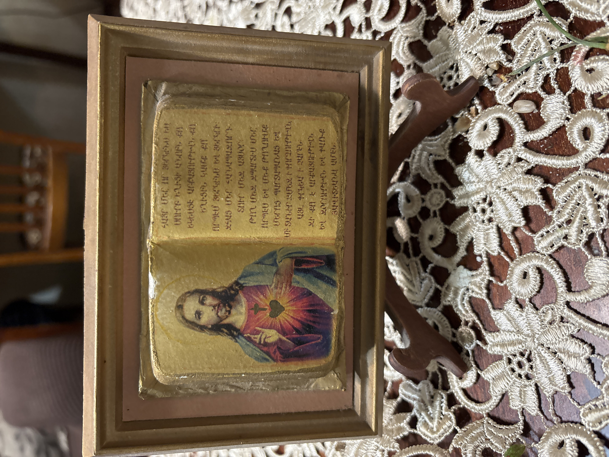 This image displays a framed devotional object, likely a religious icon or artwork, situated in Vagharshapat, Armenia. The central piece within the frame consists of two gold-toned blocks. The lower block features a printed image of Jesus Christ, depicted with his Sacred Heart radiating light and marked with a small cross; his right hand is raised. The upper block, and the area above Jesus's image, contains text written in Classical Armenian (Grabar). The visible text is identifiable as the Lord's Prayer, beginning with "ՀԱՅՐ ՄԵՐ ՈՐ ՅԵՐԿԻՆՍ" (Our Father who art in heaven), followed by the remainder of the prayer. There is also some additional faint, unclear text at the very bottom of the text block.
The devotional item is encased in a simple, light brown or gold-colored wooden frame. It rests on a dark wooden surface, which is mostly covered by an elaborate, white lace tablecloth or doily. To the right of the frame, on the lace, are scattered small, light-colored pebbles, a thin green stem, and a small, dark brown carved wooden object, possibly a decorative stand or element. The background is softly blurred, hinting at indoor furniture. The scene is illuminated by what appears to be natural daylight, suggesting a tranquil and contemplative setting.