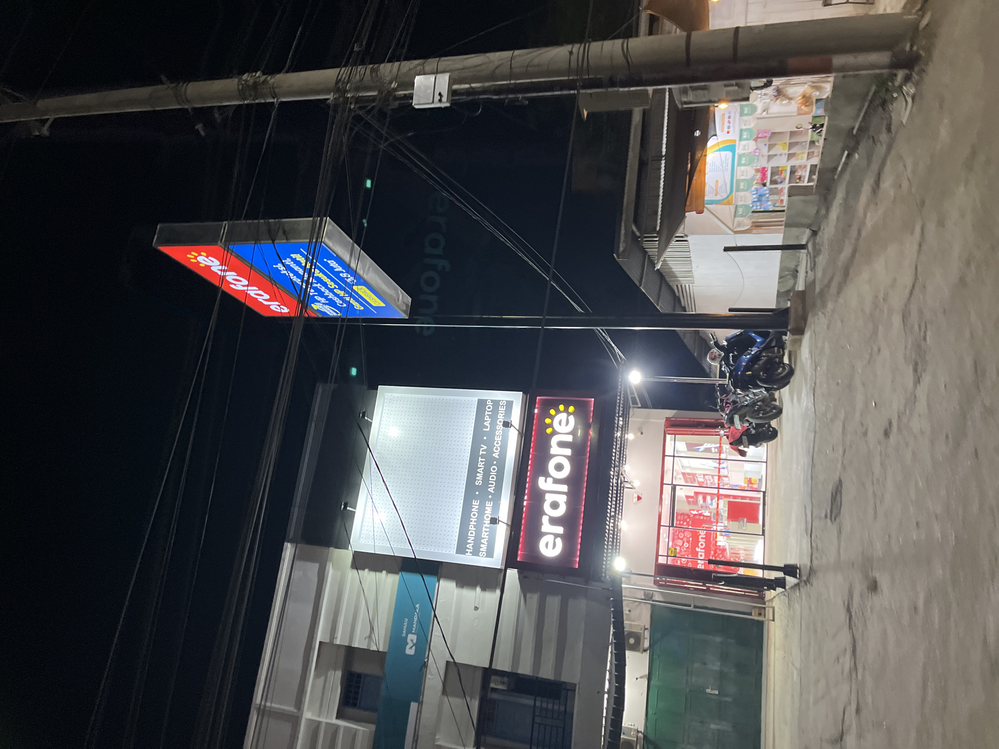 This media file depicts a nighttime scene outside a storefront in Bundar, Indonesia. The primary focus is on illuminated signage advertising "erafone," a telecommunications or electronics retailer. Multiple signs are visible, including a prominent red and blue rectangular sign with the "erafone" logo and text mentioning "HANDPHONE," "SMART TV," "LAPTOP," "SMARTHOME," and "AUDIO ACCESSORIES." Another sign, a long horizontal red one with the "erafone" logo in white, is also clearly visible.
The storefront itself appears to be modern, with large glass windows revealing shelves stocked with various products, predominantly in red packaging. In front of the store, several motorcycles are parked. The overall setting suggests a commercial area, likely a street with pedestrian and vehicle traffic, though no people are prominently featured in the foreground. The lighting indicates it is nighttime, and the concrete ground is visible, suggesting an outdoor urban environment. The image captures the vibrant, illuminated facade of the business as part of its nighttime operations. The presence of multiple wires and poles suggests overhead electrical infrastructure, common in many urban settings.