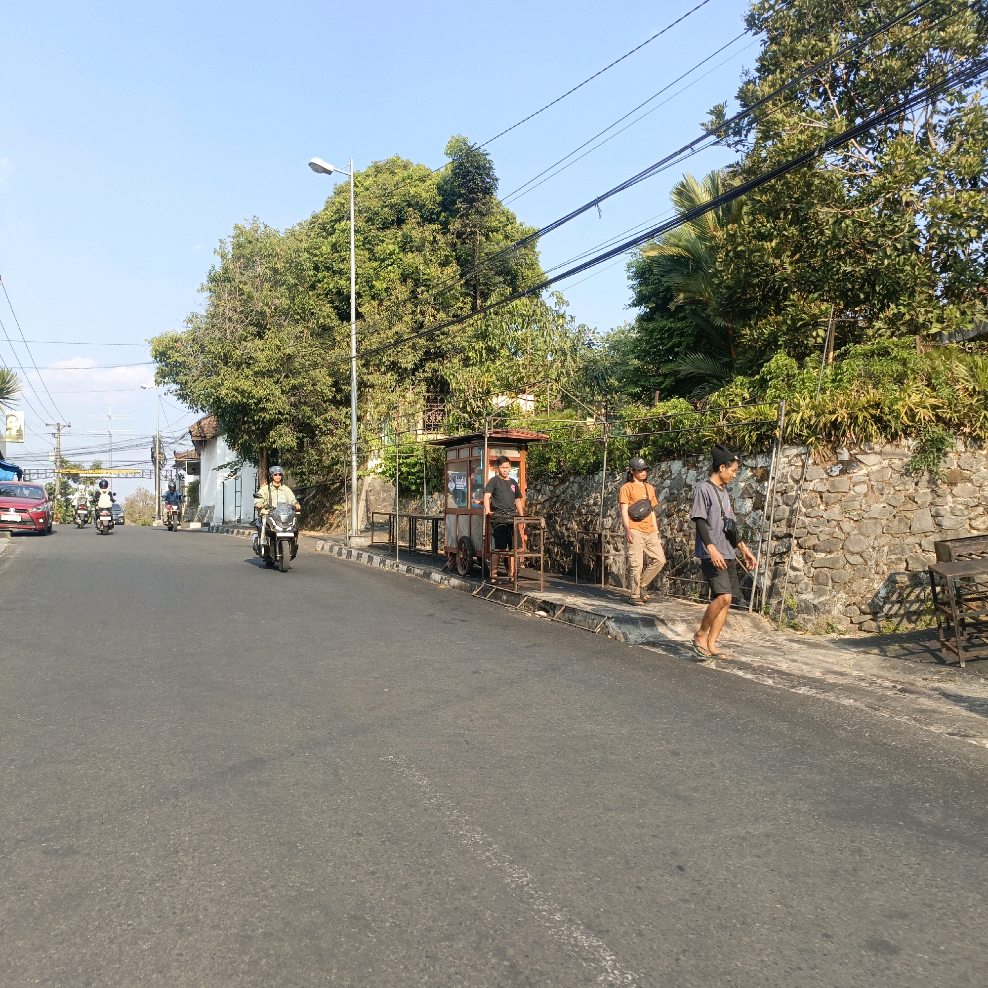 The image depicts a street scene in Salatiga, Indonesia, under a clear blue sky. A paved road occupies the foreground, with a slight incline visible. On the right side of the road, a stone wall runs along the property line, bordered by a sidewalk and a patch of green vegetation. Several people are present on the sidewalk.
On the right, two men are walking. One, wearing a light gray t-shirt, dark shorts, and a black wristband on his left arm, walks barefoot. The other, wearing an orange t-shirt, beige pants, and a black crossbody bag, is also walking.
Further down the sidewalk, closer to the road, a food cart is parked. A man in a black t-shirt and black shorts stands behind the cart. The cart itself is a mobile unit with wheels, a serving counter, and a roof.
On the left side of the road, a red car is parked. Several motorcycles are also on the road. One motorcycle, ridden by a person in a light-colored helmet and shirt, is moving away from the camera. Another motorcycle, with two riders, is further ahead, also moving away.
In the background, there are trees, buildings, and utility poles with visible electrical wires. A street lamp is present on the left side of the road. The overall impression is a sunny day in a moderately busy urban or semi-urban environment.