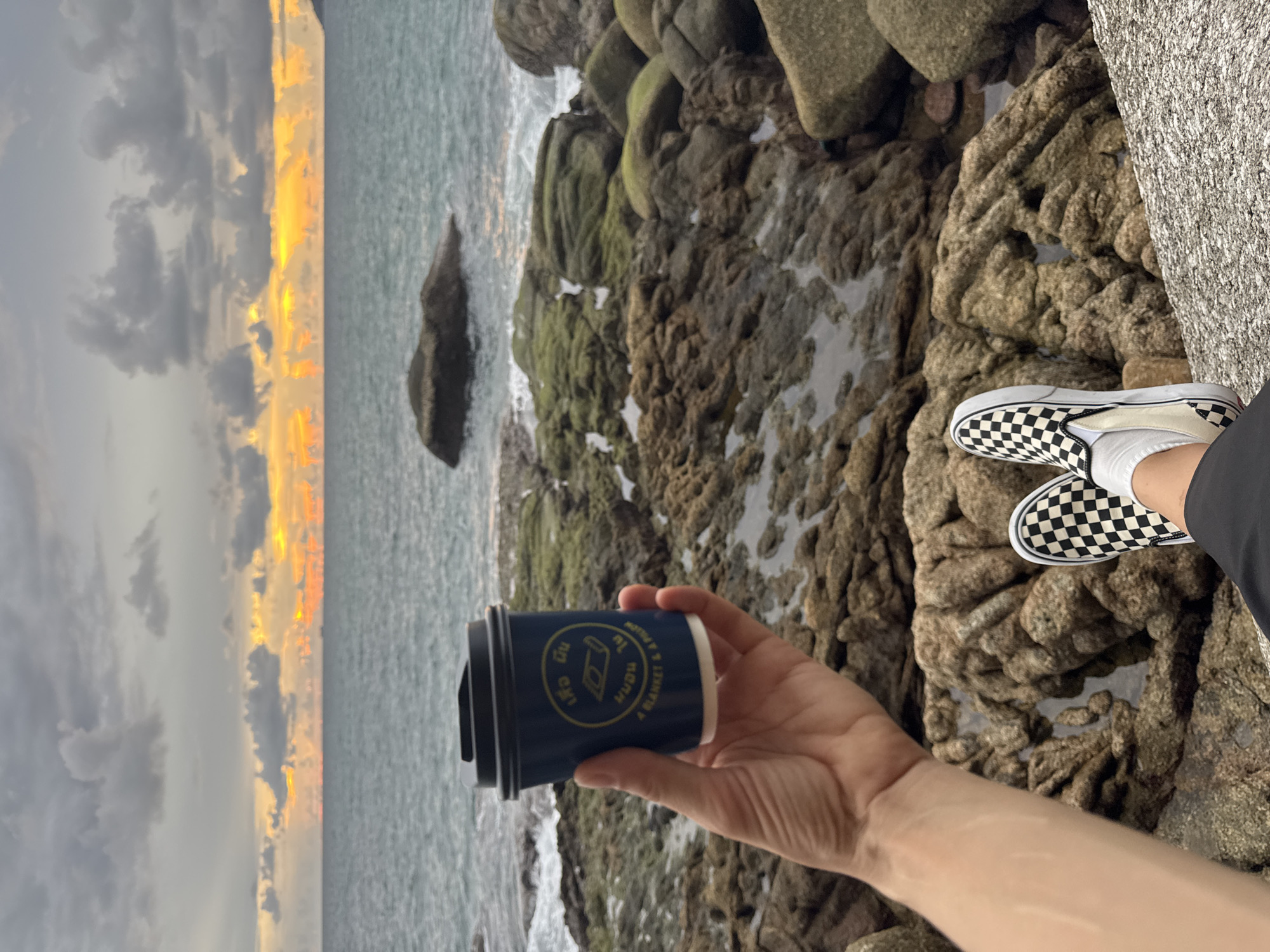 A hand holds a dark blue disposable coffee cup with yellow text and a circular logo. The cup is held in front of a rocky beach with the ocean in the background. A person's legs, wearing black and white checkered slip-on shoes and white socks, are visible at the bottom right of the frame. The sky is cloudy with a hint of sunset colors visible near the horizon. The location is identified as Karon, Thailand.