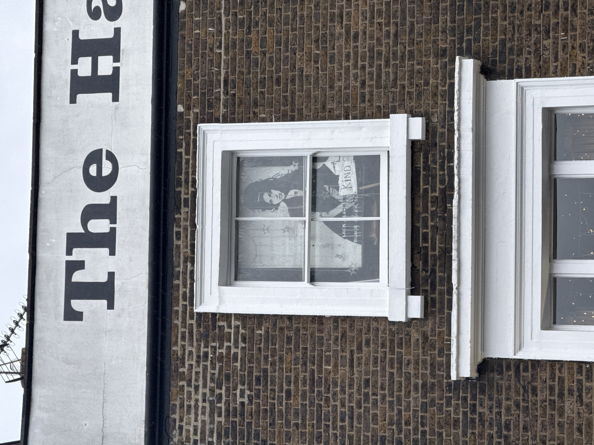 Exterior shot of a building, seemingly in London, United Kingdom, based on architectural styles. A sign reads "The Ha..." on the left side of the image. A window is visible on a brown brick wall, with a poster showing a woman and the word "KIND" visible inside. Another window is seen adjacent to it, showing interior lights.