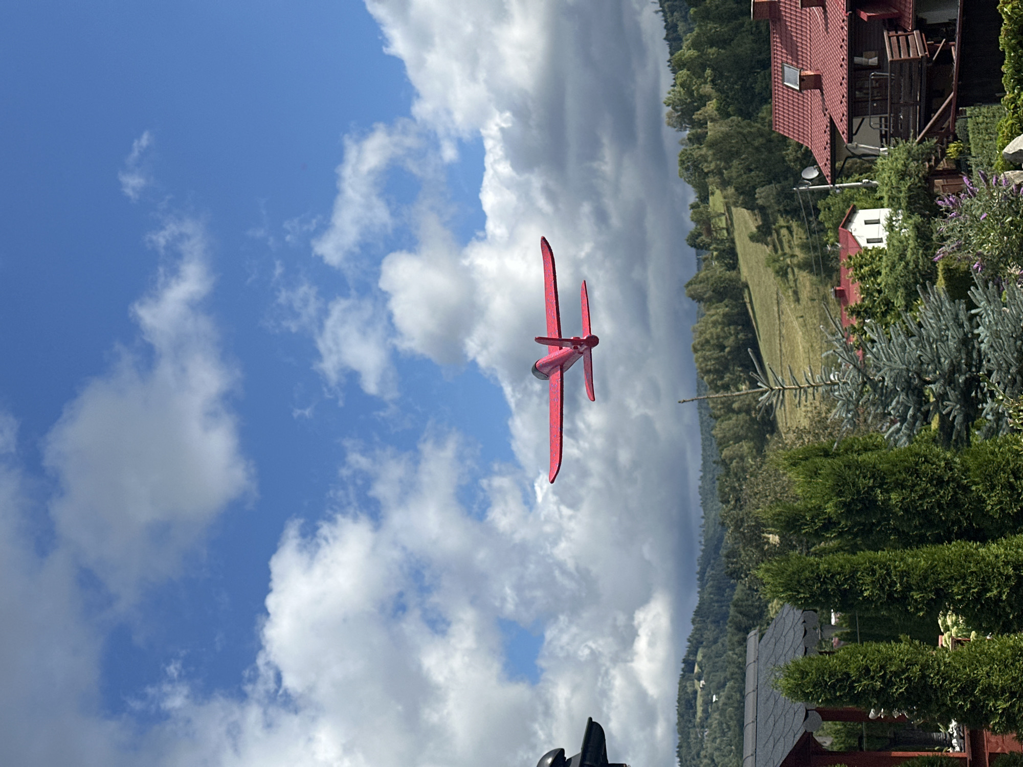 A red, model airplane is captured mid-flight against a blue sky filled with fluffy white clouds. The airplane has a single propeller at the front and two wings. In the background, rolling green hills are visible, dotted with trees and a few houses with red roofs, suggesting a rural or suburban setting. The image was taken in Wisła, Poland.