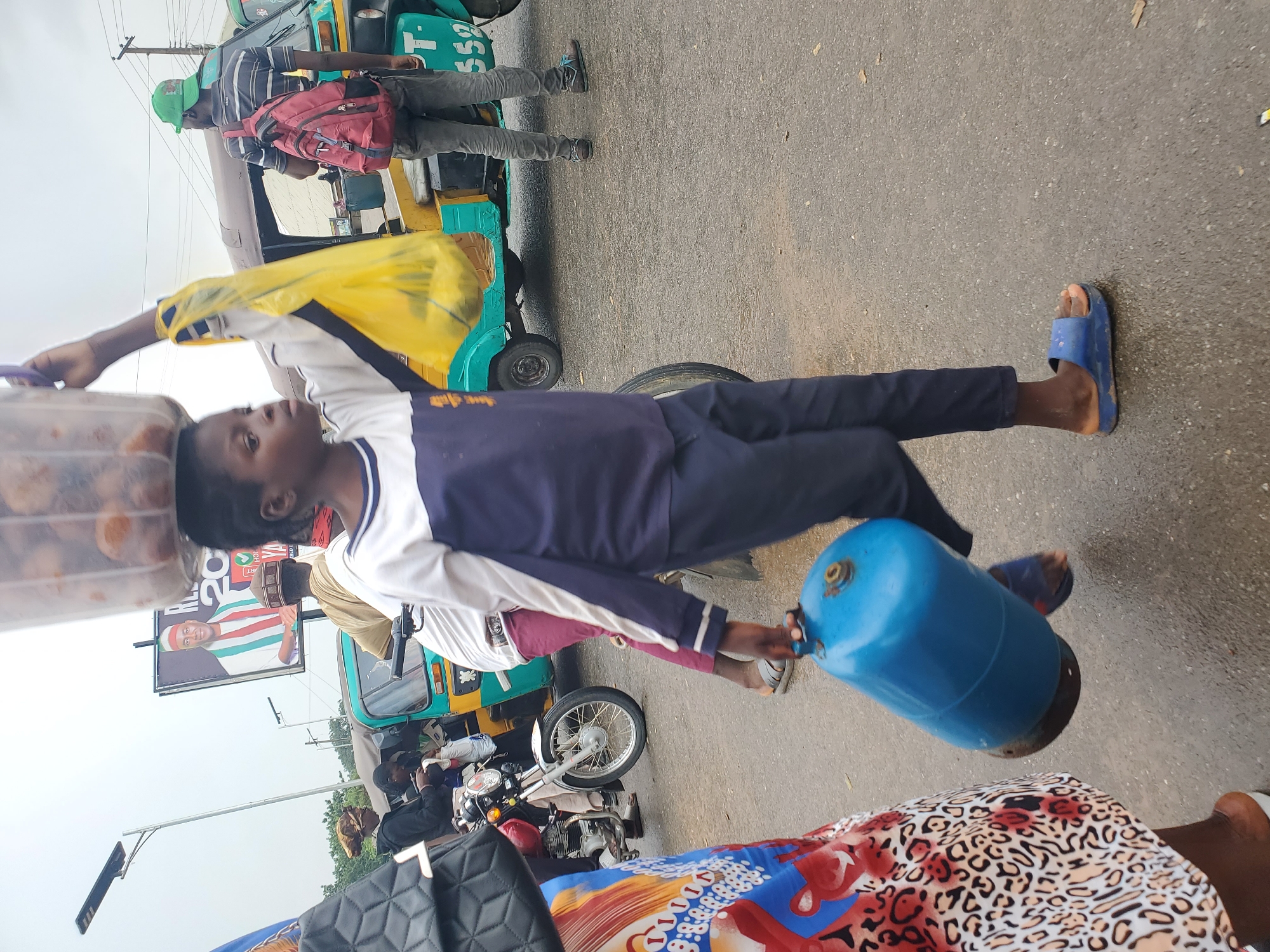 In Gwong, Nigeria, a young individual is captured mid-stride, carrying a large blue gas cylinder in their right hand and a yellow plastic bag in their left. Their head is adorned with a clear plastic container filled with what appears to be food items. They are dressed in a long-sleeved shirt with navy blue and white stripes and dark pants, along with blue slide sandals. The scene is set on a wet asphalt road, indicative of recent rain.
In the background, several vehicles are present, including a yellow and teal auto-rickshaw with the number "PT-558" visible on its side. A man wearing a green cap and a red backpack stands near the auto-rickshaw. To the left, a motorcycle is parked, and a person wearing a patterned dress walks away from the camera. Further back, a billboard displays a portrait of a man in official attire. The sky is overcast, suggesting cloudy weather. The overall atmosphere conveys a sense of daily life and commerce in a bustling urban environment.