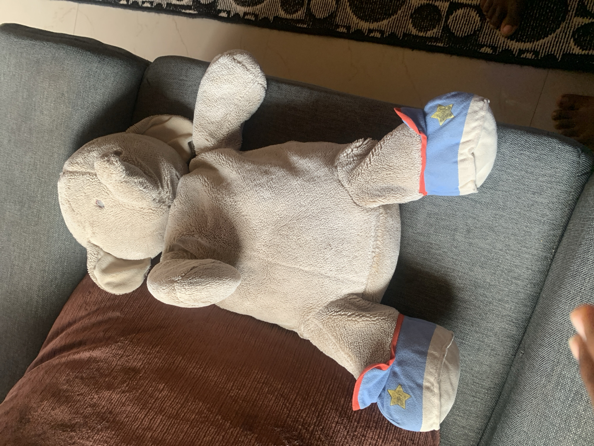 A light beige, plush toy elephant is seated on a gray sofa. The elephant is facing forward, with its legs splayed out. It wears blue booties with red trim and a yellow star on each. A rust-colored pillow is to the elephant's left. To the elephant's right, a patterned rug is visible on the floor, with a person's feet standing on it. The setting appears to be indoors in Sabon Tasha, Nigeria.