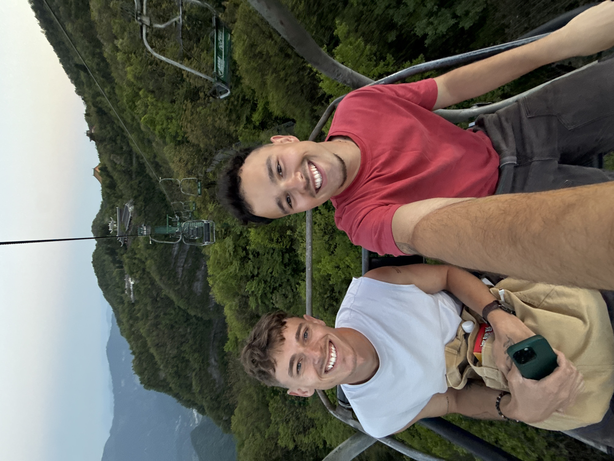 Two young men are riding a chairlift above a heavily forested mountainous area. The man on the bottom wears a white t-shirt and holds a smartphone, with a beige bag next to him. The man on top wears a red t-shirt. The chairlift extends above the treetops and toward the mountains in the distance. Other chairlifts are visible in the background. The location is in Yongding District, China.